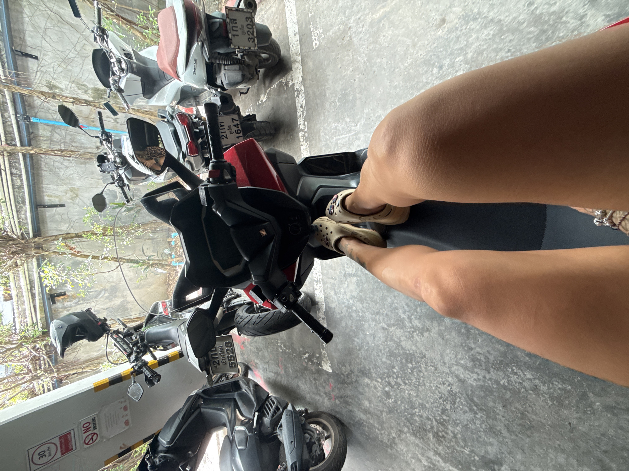 A first-person perspective shot shows a person's legs and feet, wearing tan clogs with floral embellishments, resting on the seat of a red scooter. The scooter's handlebars, a black helmet, and a portion of the red body are visible in the foreground. Behind the scooter are three other scooters parked in a lot. The ground is concrete, marked with white lines. In the background, a wall is covered in vines, and a light-colored post with signs, one indicating "30 km/hr" and another with "NO SMOKING," is visible on the left. The scene is likely in a parking area in Choeng Thale, Thailand.