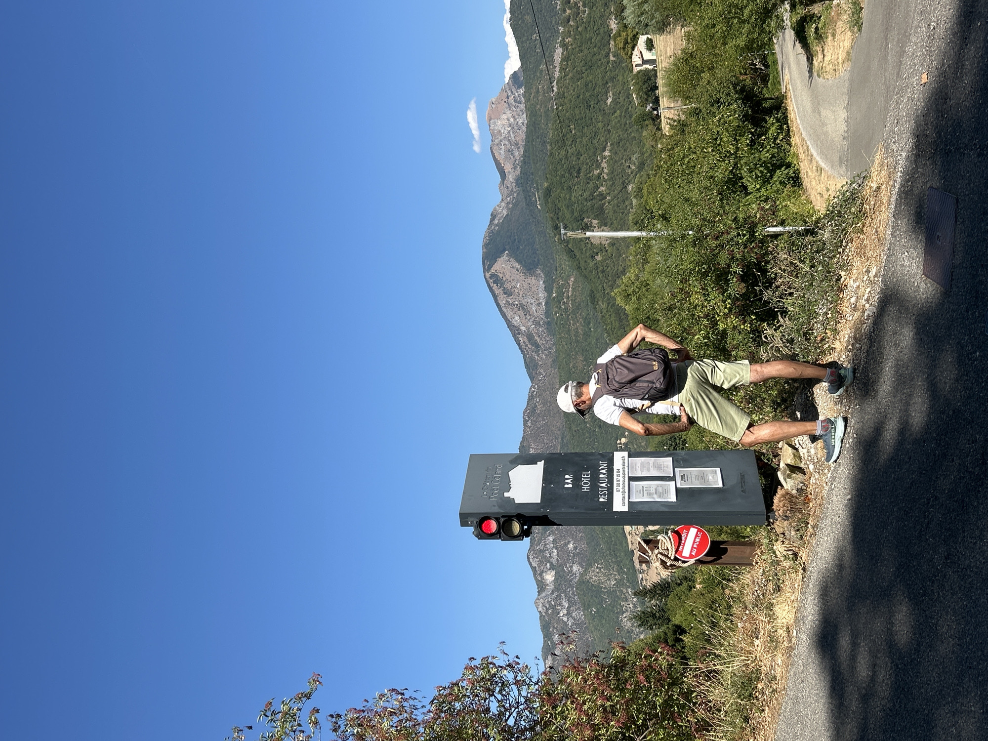 A man wearing a white baseball cap, a white t-shirt, khaki shorts, and athletic shoes stands facing away from the camera, looking at a tall black sign. The sign has a traffic light on top which is red. Below the traffic light, the sign displays "Le Château du Poët-Célard" and a graphic of a castle. Further down, it lists "BAR HÔTEL RESTAURANT" with a phone number and email address. The man has a dark backpack on.
He is standing on a paved road, next to a grassy, dry verge. To the left of the sign, a red circular sign with white text that reads "INTERDIT AU PUBLIC" (forbidden to the public) is attached to a wooden post with rope.
In the background, there are green, tree-covered hills and mountains under a clear blue sky. A winding asphalt road is visible in the middle ground, and a white building with a red roof is nestled among trees on the right side of the background. The image appears to be taken in Le Poët-Célard, France, based on the sign.