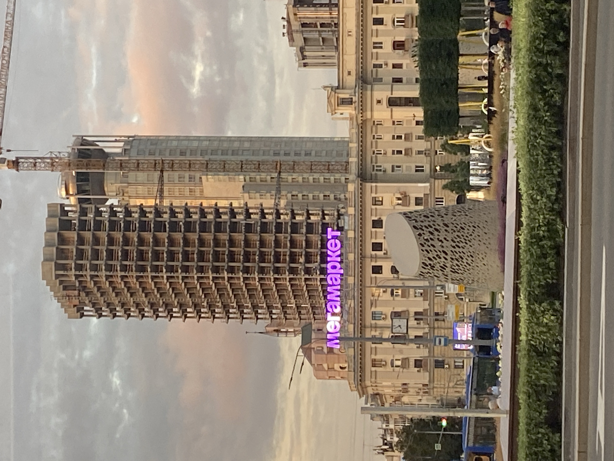 The image captures an urban scene in Moscow, Russia, focusing on a tall, unfinished skyscraper under construction. The building's skeletal structure is visible, revealing multiple floors and exposed concrete. To the left of the skyscraper, a shorter, older building with a beige facade and numerous windows stands prominently. Above this older building, a bright purple neon sign spells out "Мегамаркет" (Megamarket). In the background, other buildings and a crane suggest a developing urban landscape. In the foreground, a green hedge lines a sidewalk, and a blue bus is partially visible on the street. The sky is overcast with hints of pink from what appears to be a sunset.