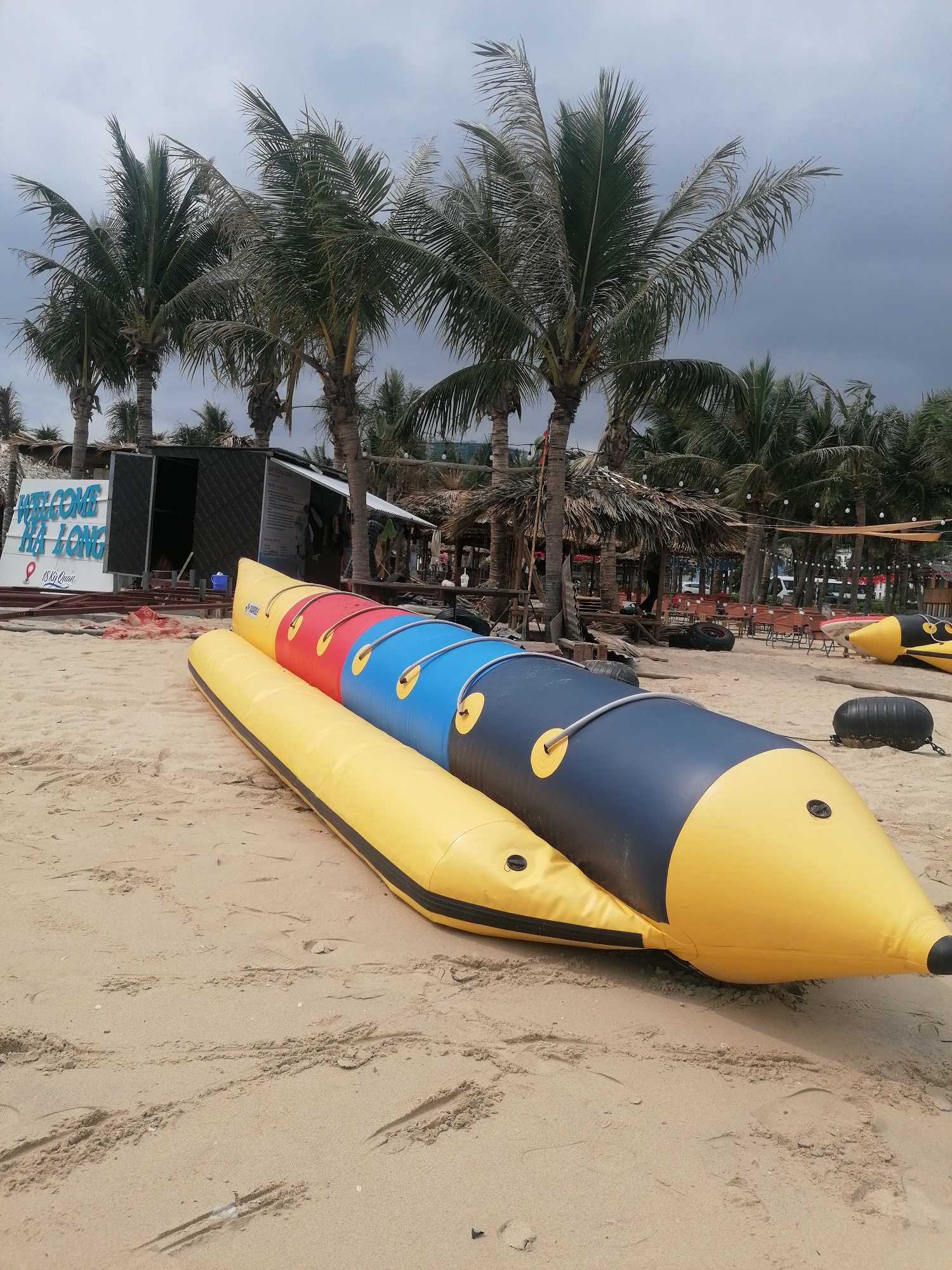 This image captures a beach scene in Bai Chay Ward, Vietnam, under an overcast sky. In the immediate foreground, a large, multi-sectioned inflatable watercraft, resembling a banana boat, lies on the sandy beach. It features segments of yellow, red, blue, and black, with a distinct yellow base running its length. Another smaller black inflatable is visible further back on the right.
Behind the main inflatable, numerous tall palm trees dominate the midground, their fronds appearing somewhat windswept against the gray sky. To the left, a prominent white sign with blue lettering reads "WELCOME HA LONG," followed by "Vila Loun" and "18 Kỳ Quan" (meaning "18 Wonders"). Adjacent to the sign is a dark-colored building or shack with open doors.
The background reveals more beachside infrastructure, including additional palm trees, thatched-roof huts, and structures that appear to be tables and chairs for a recreational area or resort. The sky is consistently cloudy and gray, indicating overcast weather during daylight hours. No people are clearly visible engaging in activities. The sandy ground shows various tracks and indentations, suggesting recent movement of objects or foot traffic. The overall atmosphere is quiet, possibly due to the cloudy weather or a lull in beach activities.