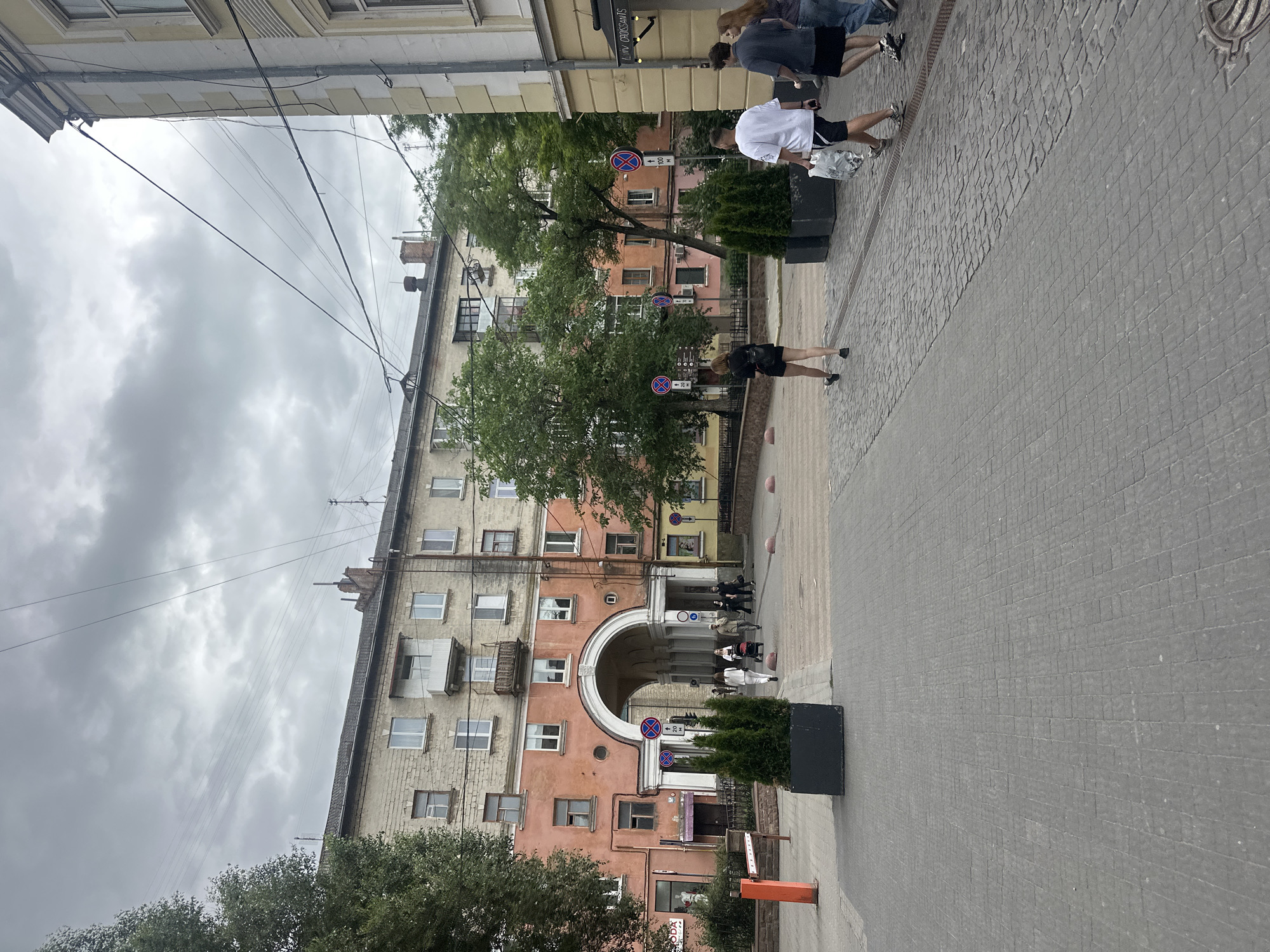 The image shows a street scene in Ternopil, Ukraine, under a cloudy sky. In the foreground, a paved walkway is visible with people walking. To the left, a large archway leads into a courtyard with buildings on either side. One building has an orange facade with multiple windows. To the right, another building with a yellow facade and architectural details lines the street. Several trees are scattered throughout the scene. There are street signs and planters with small evergreen trees. The overall impression is of a residential and commercial area in a city.