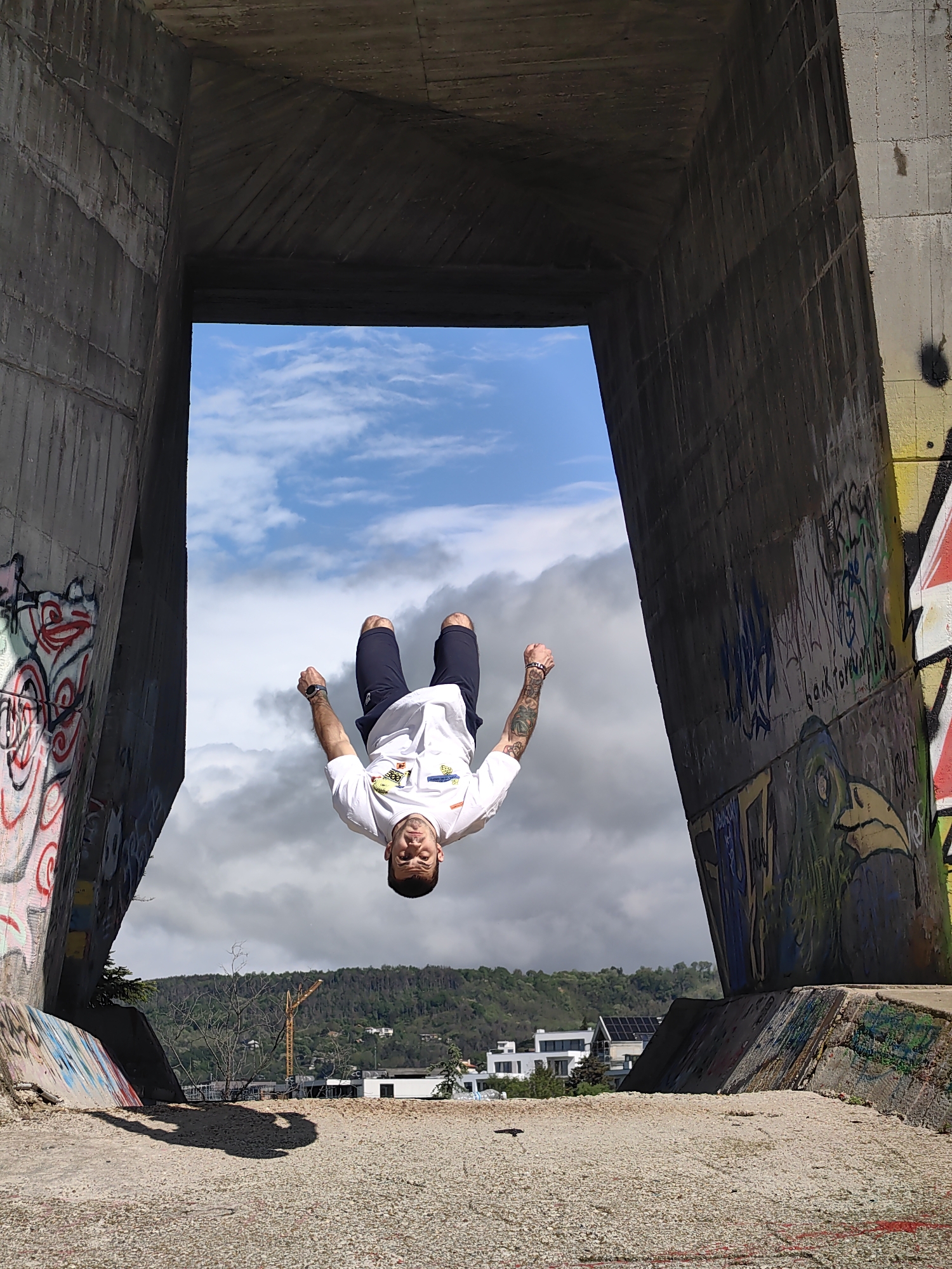 A man is captured mid-backflip, suspended upside down within a concrete structure with graffiti, against a backdrop of a partially cloudy sky and distant buildings and trees. The location appears to be an urban area, possibly Varna, Bulgaria. The graffiti on the concrete structure adds an element of urban art, and the backdrop includes buildings and a green, hilly landscape. The ground in the foreground is concrete.