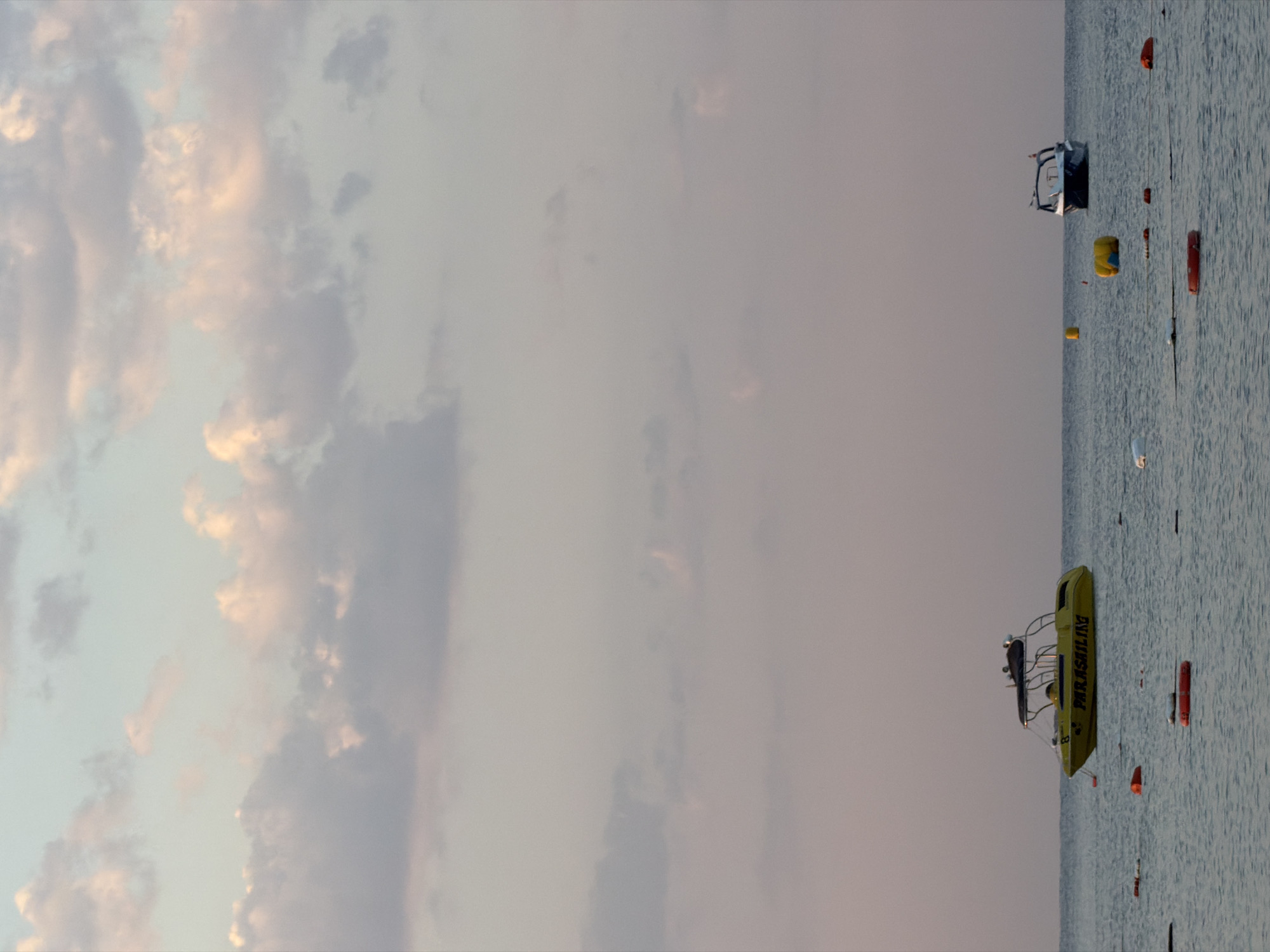 The image depicts a serene coastal scene at what appears to be sunset or sunrise, with a muted color palette dominated by soft blues, pinks, and grays in the sky and water. The sky is filled with scattered, wispy clouds that are softly illuminated, suggesting the gentle light of dawn or dusk. The sea is calm, with gentle ripples reflecting the sky's colors.
In the foreground and mid-ground, several objects are floating on the water. Most notably, a bright yellow boat with "PARASAILING" emblazoned on its side is positioned to the left of the frame. This boat appears to be a commercial vessel, possibly for recreational activities. To the right, a smaller, dark-hulled boat is visible, its silhouette distinct against the horizon. Scattered across the water are various buoys, some red and ring-shaped, others yellow and cylindrical, likely marking boundaries or hazards.
The absence of people in the immediate frame suggests a quiet moment, perhaps before or after a day's activity. The overall atmosphere is tranquil and expansive, with the vastness of the sky and sea creating a sense of peace. The location is identified as Manavgat, Turkey, suggesting this is a scene from its coastline.