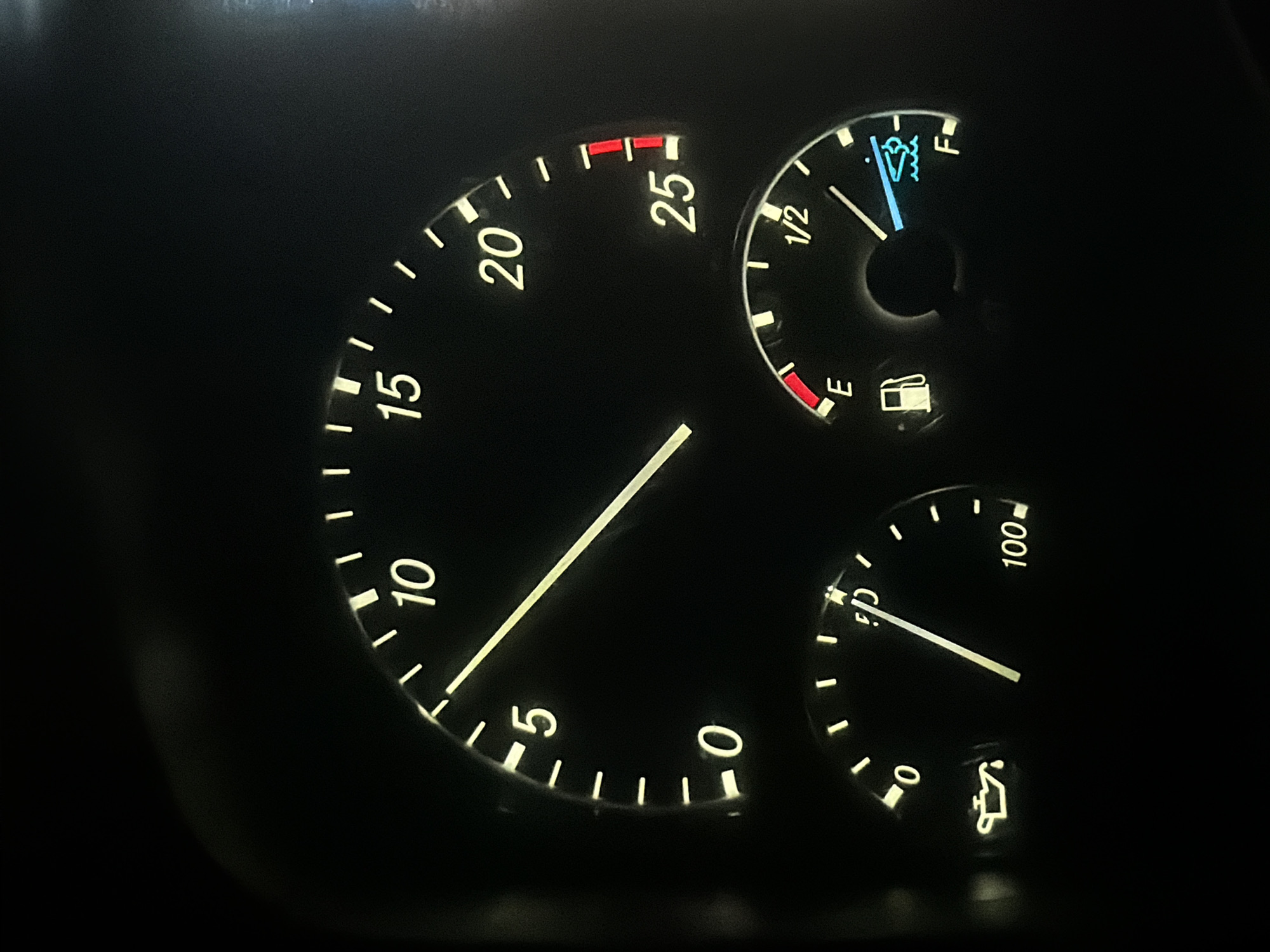 The image shows a close-up of a vehicle's dashboard at night. The dashboard features three illuminated gauges. The largest gauge, likely a speedometer, displays markings from 0 to 25, with a needle pointing slightly past the 10 mark. To the lower left, a smaller gauge, presumably an oil pressure or temperature gauge, shows markings from 0 to 100, with a needle indicating a value around 40. A symbol of an oil can is also visible near the 0 mark. To the lower right, another gauge, representing a fuel level, has markings for "E" (empty) and "F" (full), with a needle pointing past the "1/2" mark towards "F". A fuel pump icon is located near the "E" mark, and a boat icon with waves is visible near the "F" mark, possibly indicating a fuel level warning or a specific fuel type. The overall environment is dark, suggesting it is nighttime, and the gauges are lit by the vehicle's interior lighting. There are no people or other objects visible in the frame. This scene is not specifically located in Wildwood, United States; it is a generic image of a vehicle's dashboard.