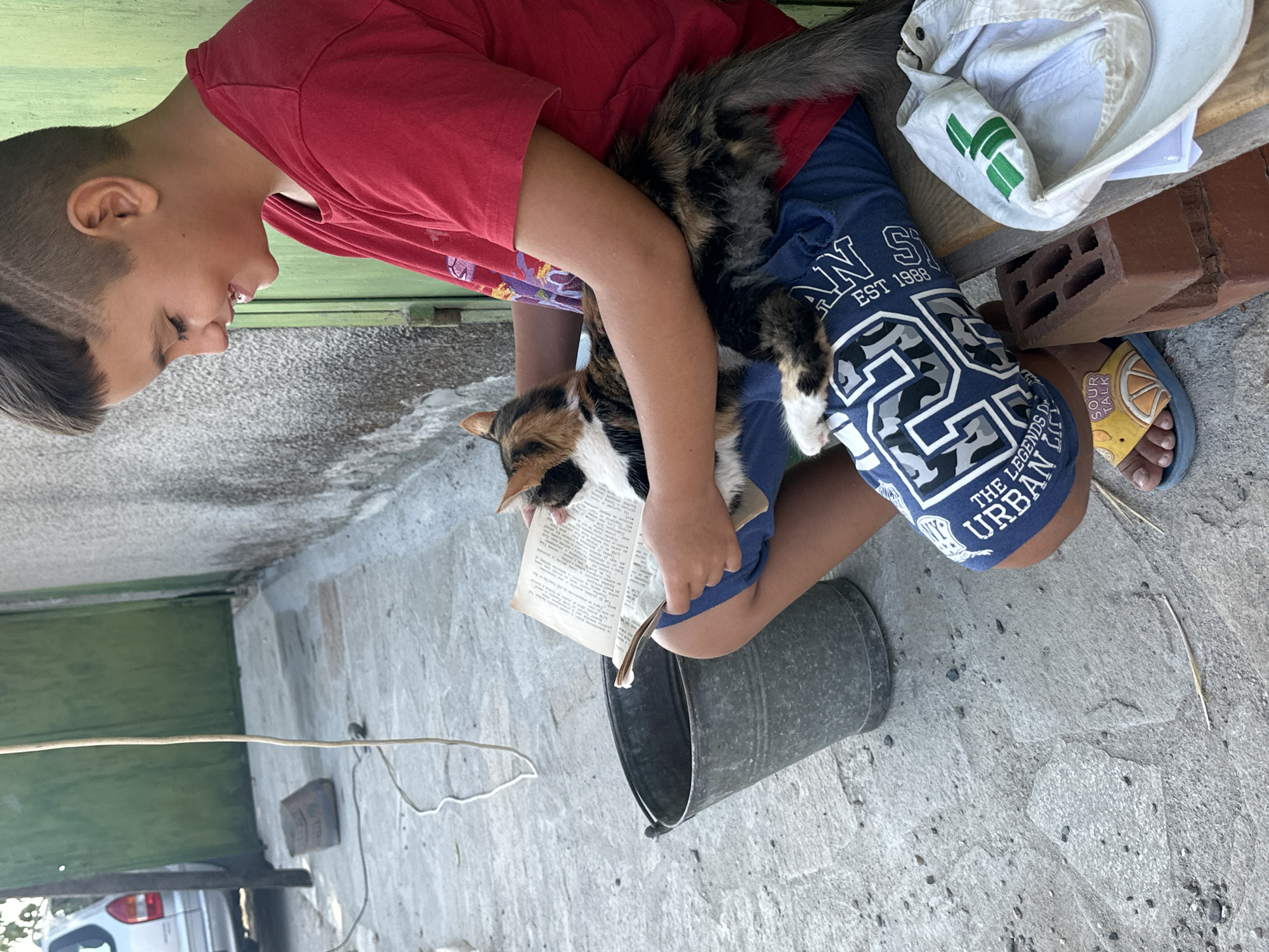 In Malevo, Bulgaria, a young boy with short, styled brown hair and a red t-shirt sits on a wooden bench, holding an open book and a calico cat. The cat is nestled on his lap, its tail extending over his blue shorts. The boy is smiling, looking down at the book. To his left is a weathered metal bucket. The ground beneath them is paved with large, irregular stones. Behind the boy, a textured grey wall meets a green wooden door. In the background, a silver car is partially visible. A white cap with a green logo rests on some red bricks to the right of the bench, near the boy's feet.