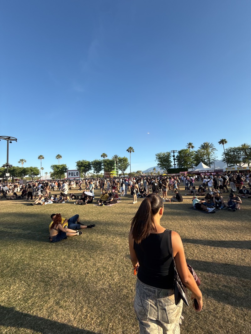 The image shows a daytime outdoor scene at the Coachella Valley Music and Arts Festival in Indio, United States. A woman with dark hair pulled into a ponytail stands in the foreground, facing a large crowd of people on a grassy field. The crowd appears to be relaxing and socializing. Some people are seated or lying on the grass, while others are standing and walking around. In the background, there are palm trees, stages, and tents. A sign reading "POLO" is visible. The sky is clear and blue.