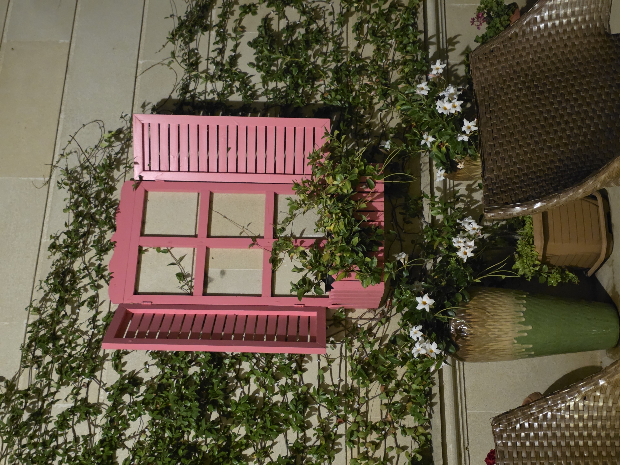 The image shows a decorative pink window box mounted on a wall in Buzovna, Azerbaijan. The window box is adorned with lush green vines and white flowers. To the left of the window box is a large, green and brown ceramic vase, and to the right, a wicker chair. The wall itself is made of light-colored stone blocks and is partially covered with climbing ivy. In the foreground, another wicker chair is visible, along with a wooden planter box. The scene appears to be an outdoor setting, possibly a patio or garden.