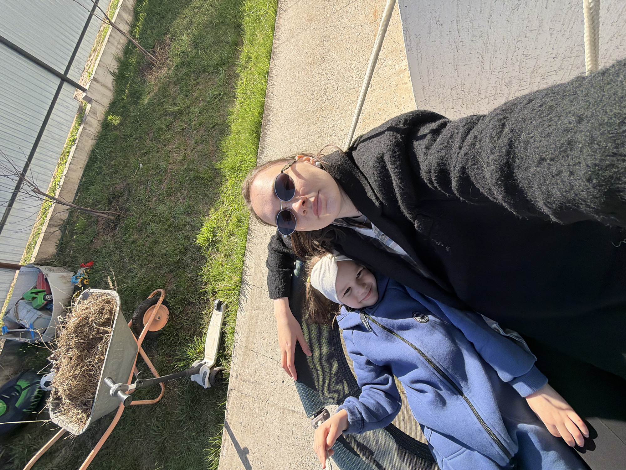 The image depicts two individuals, likely a woman and a child, positioned on an outdoor trampoline. The woman is wearing sunglasses and a dark-colored jacket or coat. The child is wearing a blue jumpsuit with a white headband. The background includes a grassy area, a concrete pathway or patio, and a section of fencing. To the left, there is a wheelbarrow filled with dried plant matter or hay, along with other unidentified objects. The location is speculated to be Prikamskii, Russia. There is a scooter visible next to the concrete pathway and grass. The woman appears to be taking a selfie or photo of herself and the child.