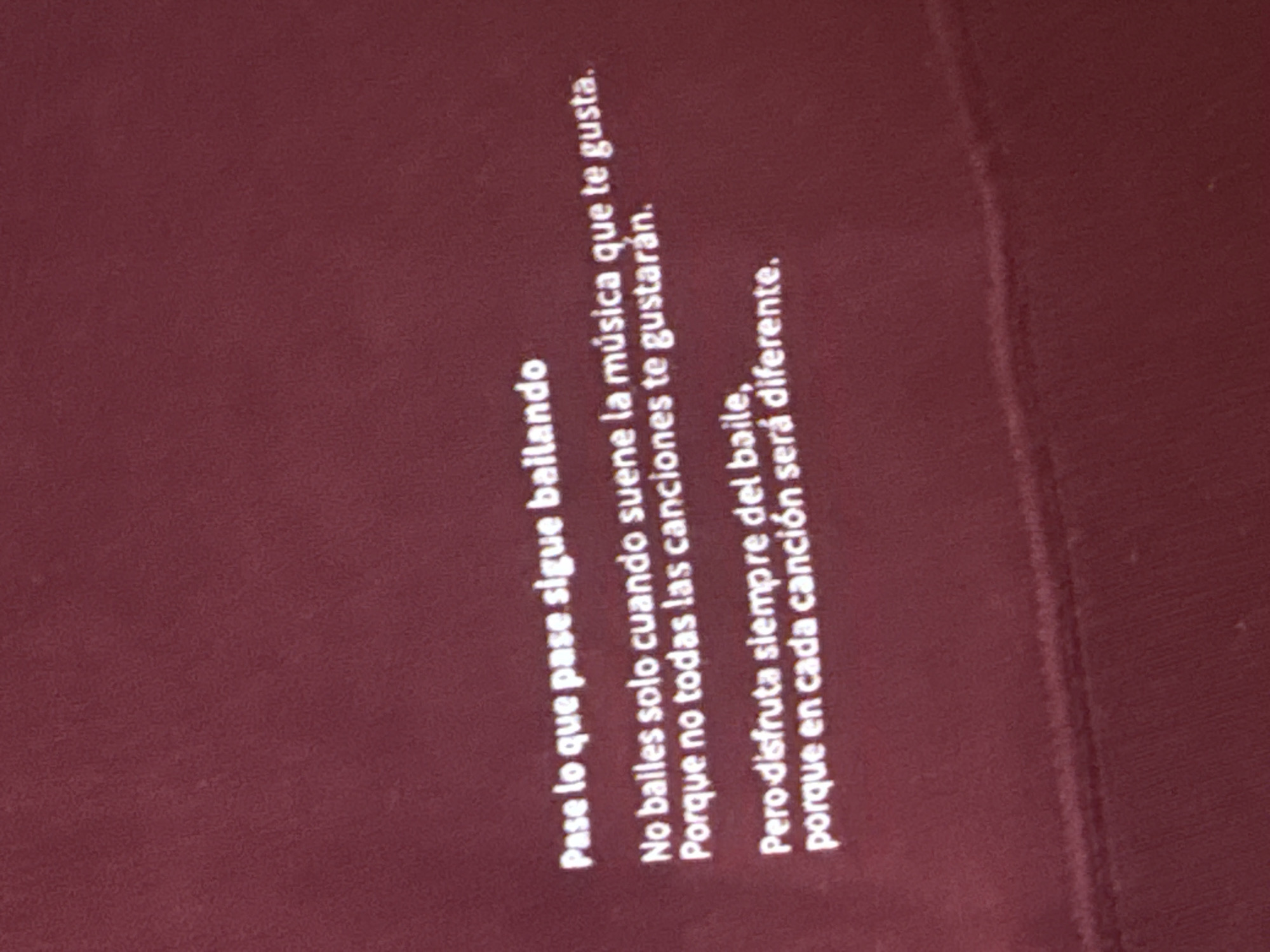 The image shows white text printed on a dark red background. The text, which appears to be in Spanish, consists of multiple lines:
"Pase lo que pase sigue bailando"
"No bailes solo cuando suene la música que te gusta."
"Porque no todas las canciones te gustarán."
"Pero disfruta siempre del baile,
porque en cada canción será diferente."
The text is vertically oriented in the image, reading from top to bottom. The background is a uniform dark red color with a slightly textured appearance. There are no people or specific objects visible, only the printed text.