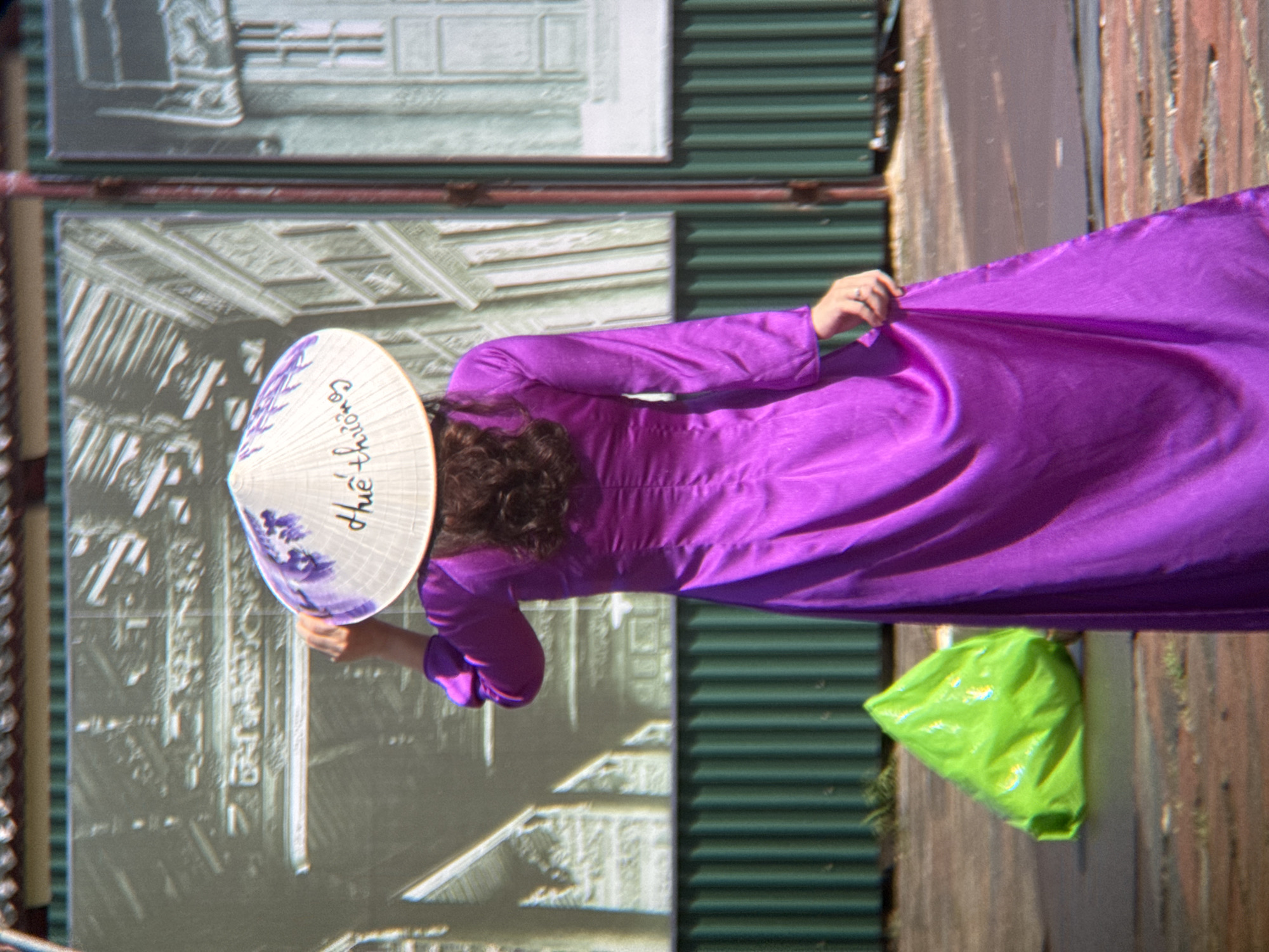 A person wearing a vibrant purple dress and a conical hat stands with their back to the camera in an outdoor setting. The hat is adorned with purple writing and a floral pattern. The dress has long sleeves and a fitted bodice with ruched detailing on the back. In the foreground, to the left, a bright green, crinkled plastic bag lies on the ground. The background features a textured wall with what appears to be a mural or large photograph depicting architectural elements, possibly from Phú Xuân Ward, Vietnam. A corrugated green fence runs across the middle ground. The ground surface is a mix of brickwork and asphalt.