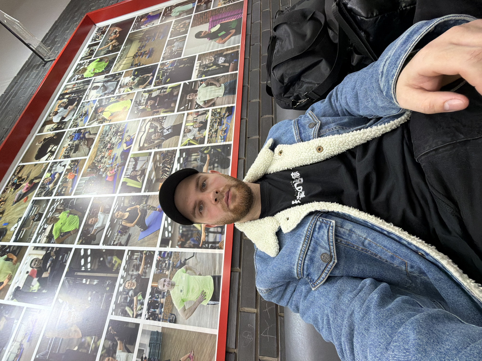 A medium shot depicts a Caucasian man with a short beard, wearing a black cap and a blue denim jacket with a white fleece lining, sitting in what appears to be the city of Chaikovsky, Russia. He is dressed in a black t-shirt with white writing. Beside him is a black duffel bag, and he is seated against a brick wall with graffiti markings. Behind him is a large wall display featuring a collage of photographs, many appearing to be people engaging in physical activities. The photos are individually framed with white borders and the entire display is outlined by a red frame.