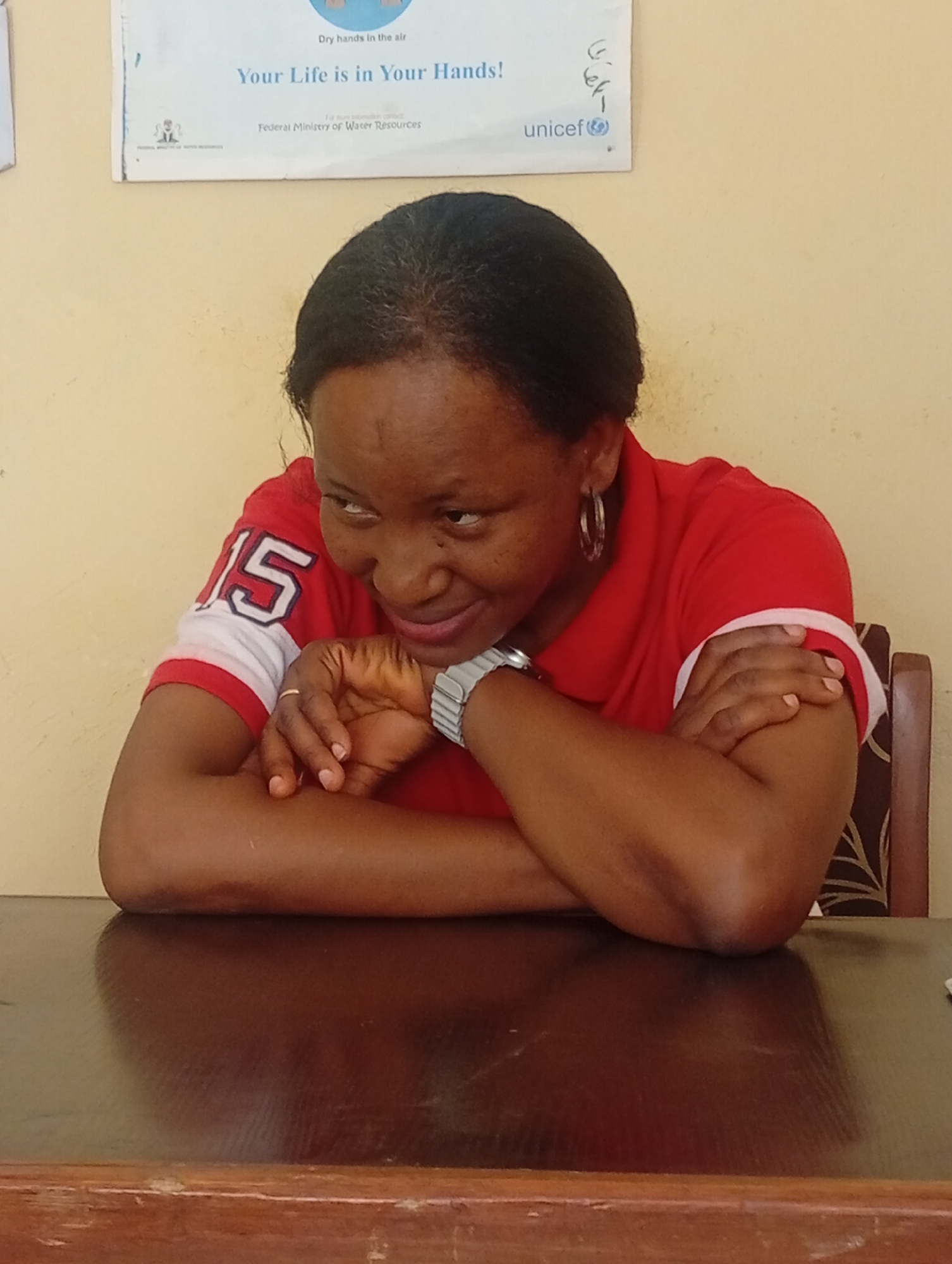 A middle-aged woman is seated at a dark wooden table, leaning forward with her arms crossed and resting on the table's surface. She wears a red polo shirt with the number "15" printed on the upper left arm, and a silver wristwatch is visible on her left wrist. Her face is slightly turned to the left, with a gentle smile. She has dark hair pulled back and is wearing hoop earrings. Behind her and slightly above, a poster is affixed to the wall, reading "Dry hands in the air" and "Your Life is in Your Hands!". Beneath this, the words "Federal Ministry of Water Resources" are printed. The UNICEF logo is visible to the right of the poster, along with illegible handwritten marks. The environment seems to be indoors with a plain, light-yellow wall serving as the backdrop.