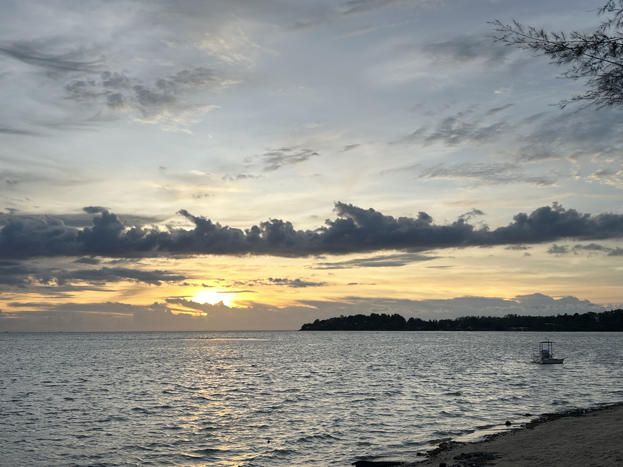 The image depicts a serene sunset over the ocean in Ko Pha Ngan, Thailand. The sky is a canvas of soft greys, whites, and warm oranges, with clouds scattered across the horizon. The sun is partially obscured by clouds, casting a golden glow on the water. The ocean is calm, with gentle ripples reflecting the sunset's hues. On the right side of the frame, a small white boat with a canopy is anchored in the shallow water near the shore. The shoreline is sandy, with some debris and a few pieces of driftwood visible. In the distance, a silhouetted island with lush vegetation marks the horizon. The overall atmosphere is tranquil and picturesque.