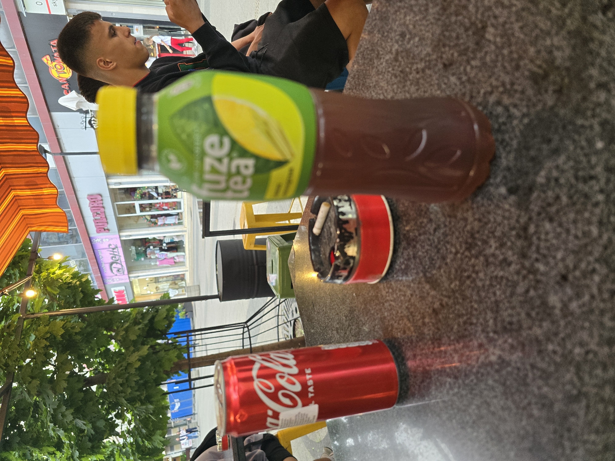 The image shows an outdoor cafe scene, likely in Pleven, Bulgaria. A young man is seated, wearing a black t-shirt and shorts, facing toward the building across the street. On the table are a bottle of Fuze Tea, a red Coca-Cola can, and a red ashtray with cigarette butts. The cafe has yellow chairs and black tables. In the background are several shops with signs in Cyrillic. Trees with green leaves are visible to the left of the scene. The image is slightly blurry and taken from an elevated perspective.