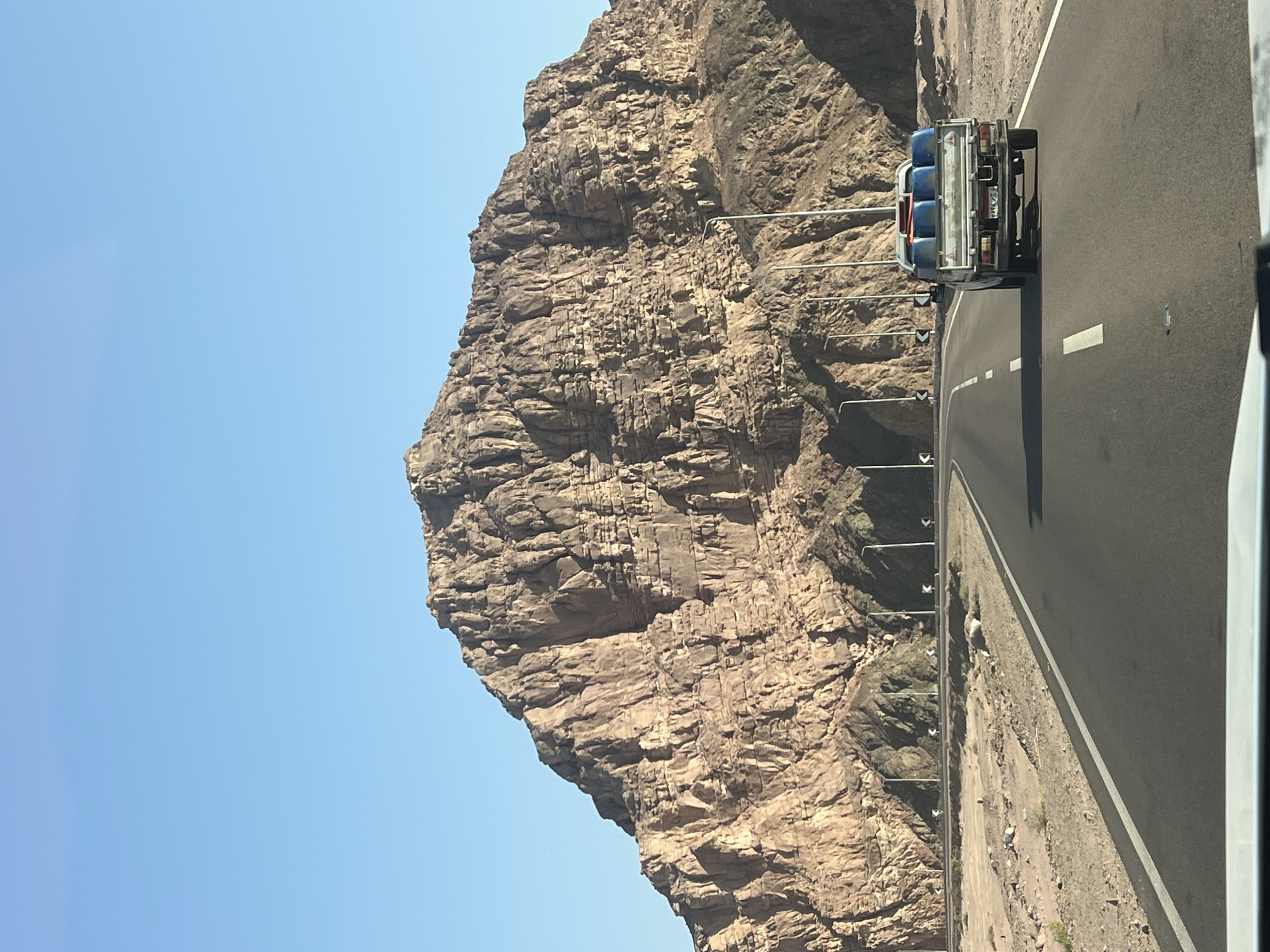 The image captures a desert landscape near Dahab, Egypt, dominated by a massive, rugged mountain bathed in sunlight. A paved road, with white lane markings and reflective markers, cuts through the arid terrain, leading towards the mountain. The road is lined with streetlights, suggesting it is a route within or leading to a populated area. In the foreground, a white pickup truck is driving away from the viewer on the right side of the road. The truck's bed is filled with blue barrels, secured with straps. The sky is a clear, bright blue. The scene is characterized by its vastness, dry environment, and the imposing presence of the rocky mountain.