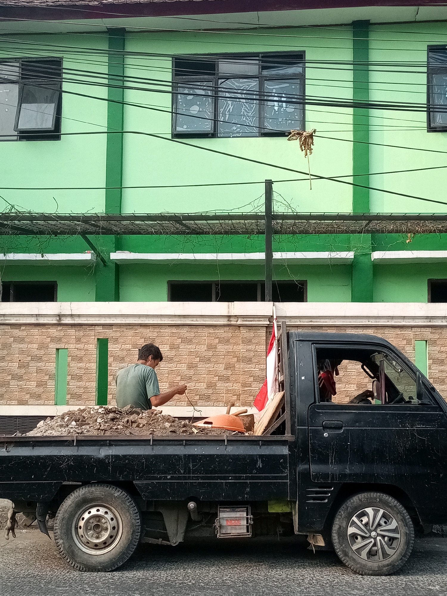 A man wearing a green t-shirt is sitting in the bed of a black pickup truck, which is filled with dirt and debris. He is holding a thin, brown string in his right hand. In the background, a light green building with dark-framed windows is visible, along with some electrical wires. A brick wall runs horizontally across the middle of the image, separating the building from the truck. A small Indonesian flag is mounted on a wooden plank in the truck bed. The truck is parked on a paved road. The overall scene suggests a construction or demolition site, likely in a residential or commercial area of Bekasi, Indonesia, given the building style and flag.