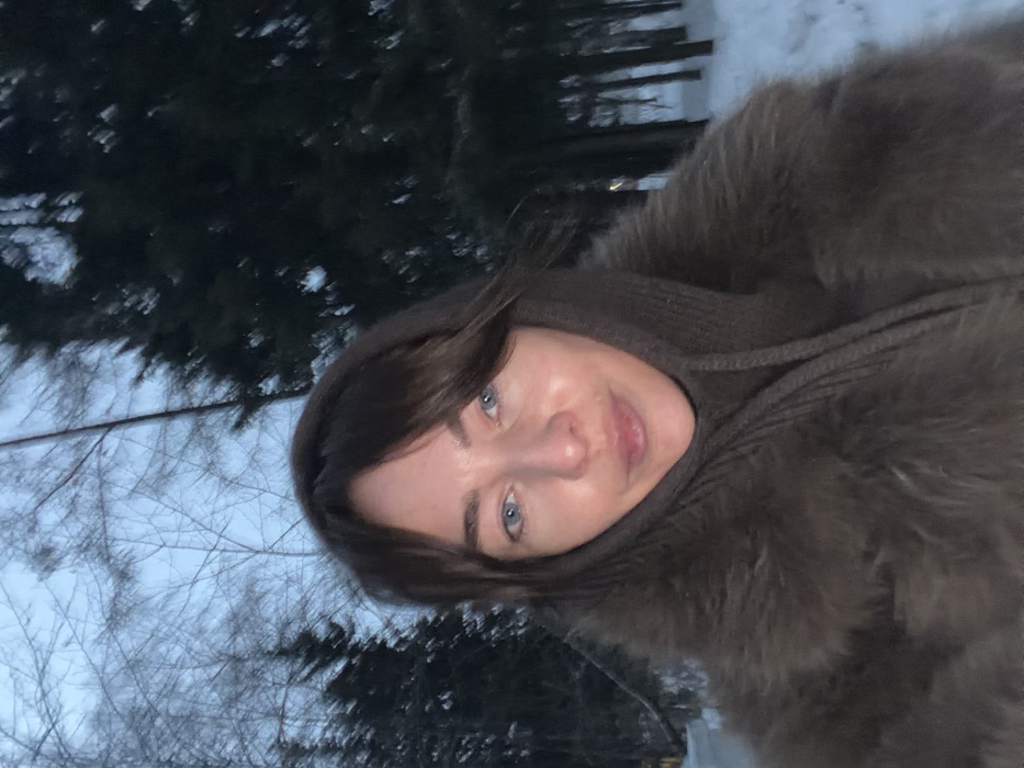 The image displays a head-and-shoulders shot of an adult female, centrally positioned, looking directly at the camera. The scene is set outdoors in a snow-covered, wooded environment in Ust-Ilimsk, Russia. The female presents with brown hair featuring a fringe, blue eyes, and fair skin. She is dressed in a dark brown garment, consistent with a fur or faux fur coat, layered over a ribbed dark brown knit balaclava or cowl covering her head and neck. The background features white snow covering the ground. To the left, bare deciduous trees with a dense network of branches are visible. To the right and behind the subject, dark green evergreen trees stand against a muted, light blue-grey sky, indicative of overcast conditions or low light levels typical of winter dawn or dusk.