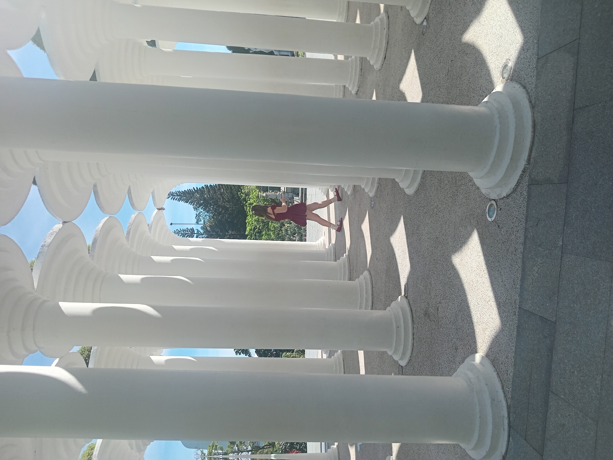 A young woman in a short maroon dress and red shoes walks through an outdoor installation of tall, white, fluted columns. The columns are arranged in rows, creating a visual effect of depth and perspective. Sunlight streams through the spaces between the columns, casting distinct geometric shadows on the speckled gray ground. In the background, lush green trees and a bright blue sky are visible. This installation is located at The Hidden Gem Da Nang, a popular tourist attraction in Da Nang, Vietnam, known for its artistic design and picturesque setting.