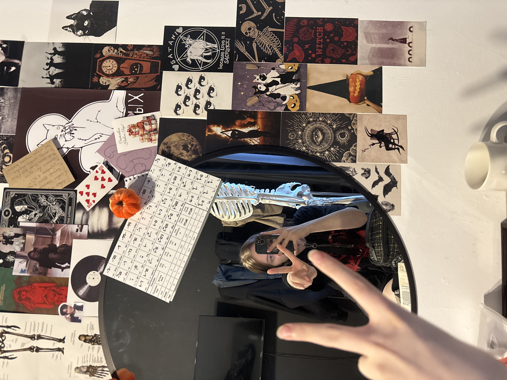 The image is an indoor selfie taken in a room with Halloween-themed decorations. A person is reflected in a round, black mirror on a black table. The person, presumably female, is holding a phone with a triple-lens camera and making a peace sign. In the reflection, there is a skeleton hanging behind the person. The table also has a calendar written in Thai, a small orange pumpkin, vinyl record, and cards. The wall behind the table is covered with Halloween-themed images and stickers, including cats, skeletons, and witches. A white mug is visible in the upper right corner. The overall ambiance suggests a focus on alternative themes. There is no indication this photo was taken in Верхнеказымский, Russia.