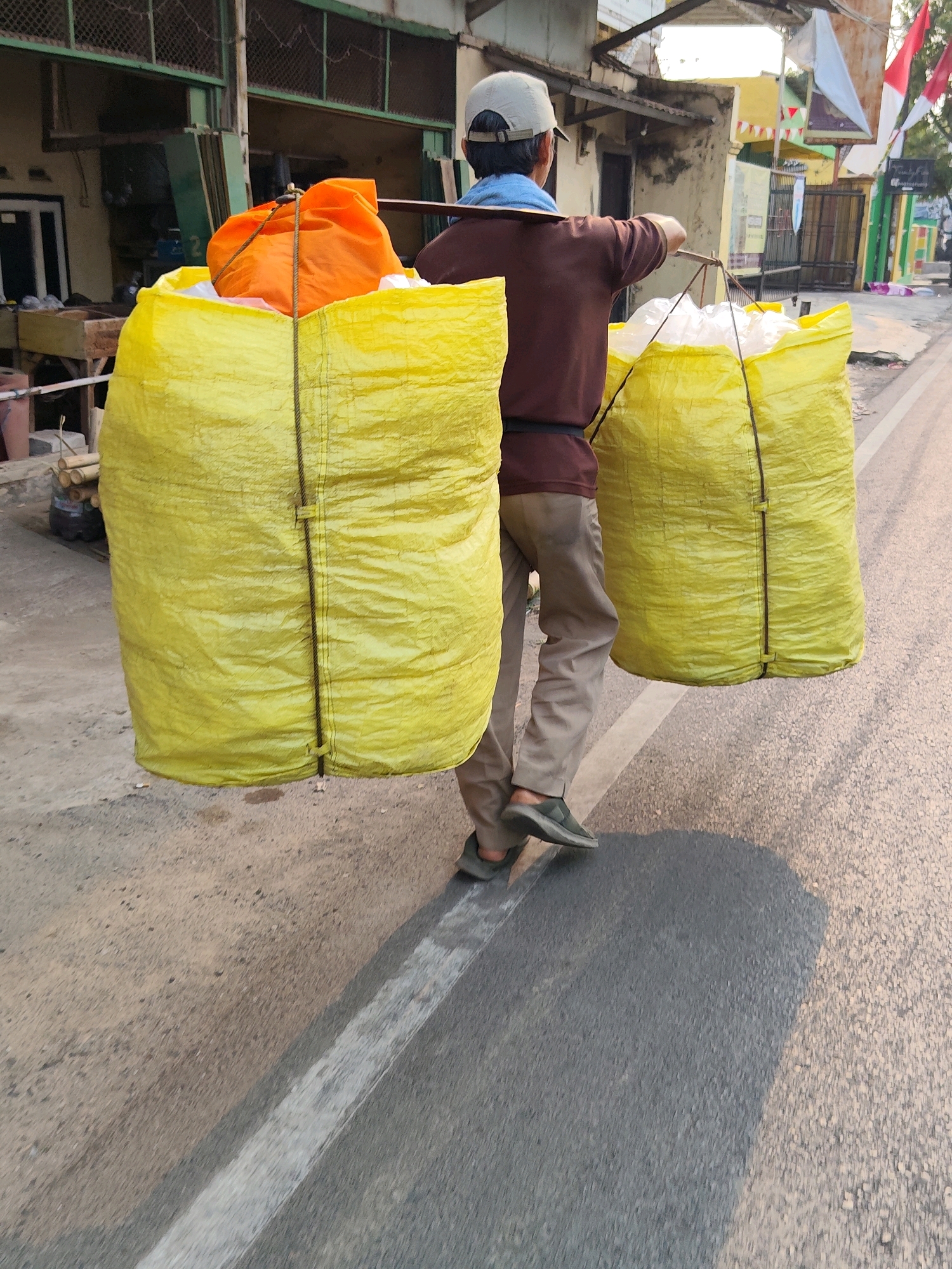 A person, from behind, wearing a brown long-sleeved shirt, khaki pants, a beige baseball cap, and dark sneakers, walks on a street in Purwakarta, Indonesia. They are carrying two large, bright yellow sacks filled with what appears to be trash. The sacks are slung over a wooden pole resting on their shoulders. Inside the left sack, a bright orange bag is visible, also filled with contents. The street is asphalt, with a white line marking the lane. On the right side of the street, there are buildings with corrugated metal roofs and some decorative flags, one of which appears to be the Indonesian flag. The scene is illuminated by natural light, suggesting it is daytime.