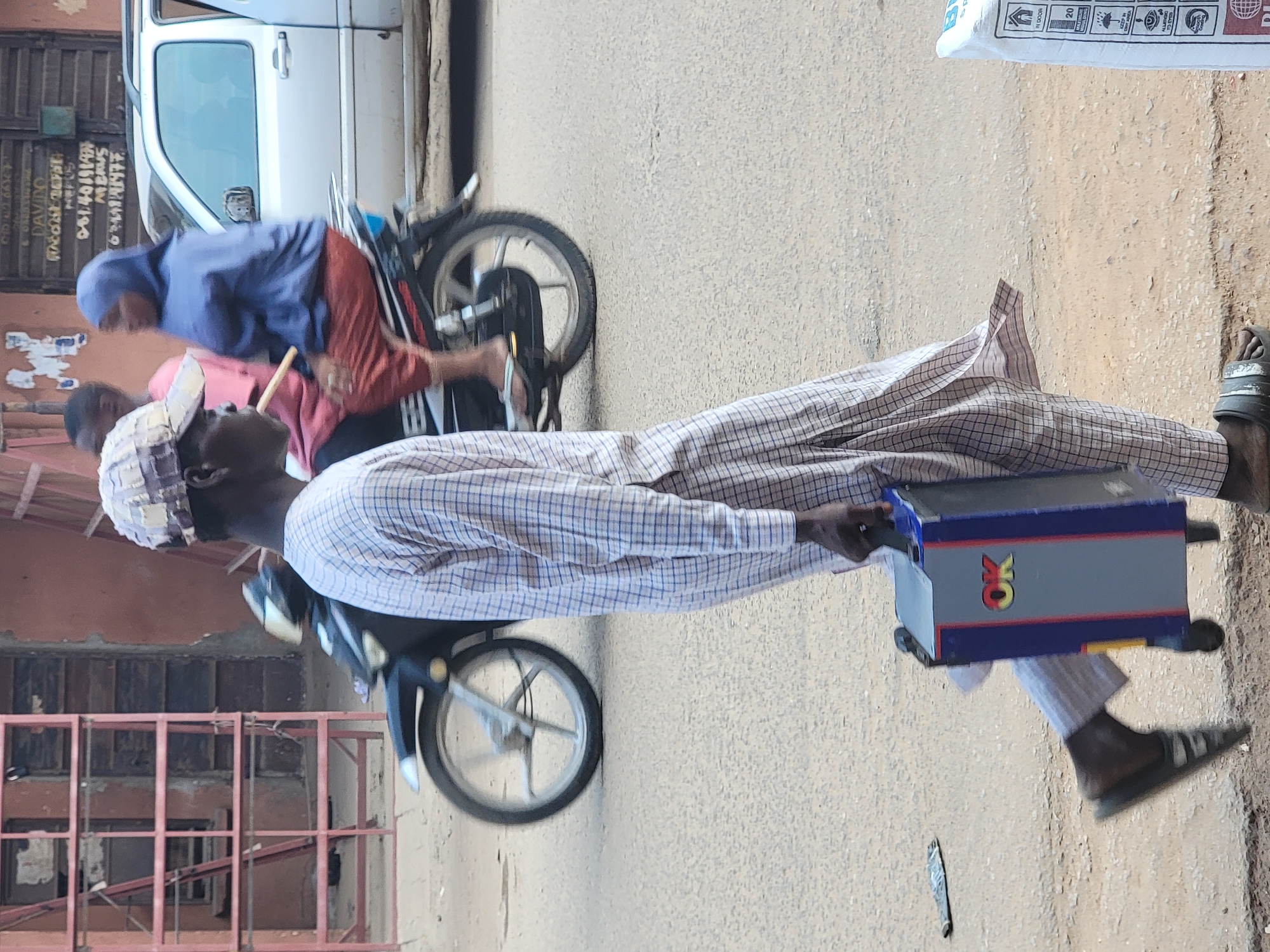 In Maiburiji, Nigeria, an adult male dressed in a plaid shirt, pants, and a patterned hat carries a blue and gray box labeled "OK." He walks along a paved road in front of a building under construction. Adjacent to him, a man and a woman on a motorcycle face the opposite direction. A white vehicle is parked nearby.