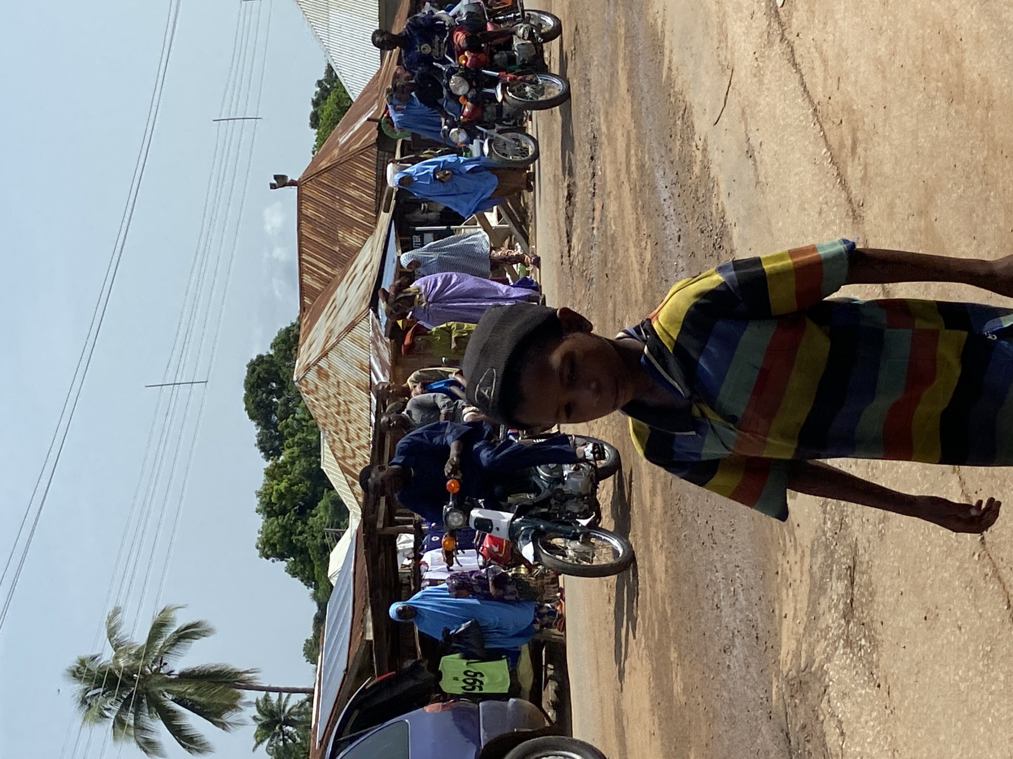 A medium, ground-level shot depicts a street scene in Mararaba, Nigeria. A boy wearing a striped shirt and a black kufi cap stands on the right side of the frame. To the left, a line of people is seen on motorbikes, some wearing traditional African attire, under a corrugated metal roof, suggesting a market area. A dark-colored car is partially visible at the bottom left of the frame, and a palm tree stands in the background. Overhead electrical wires are seen against a light blue sky. The road appears to be unpaved.