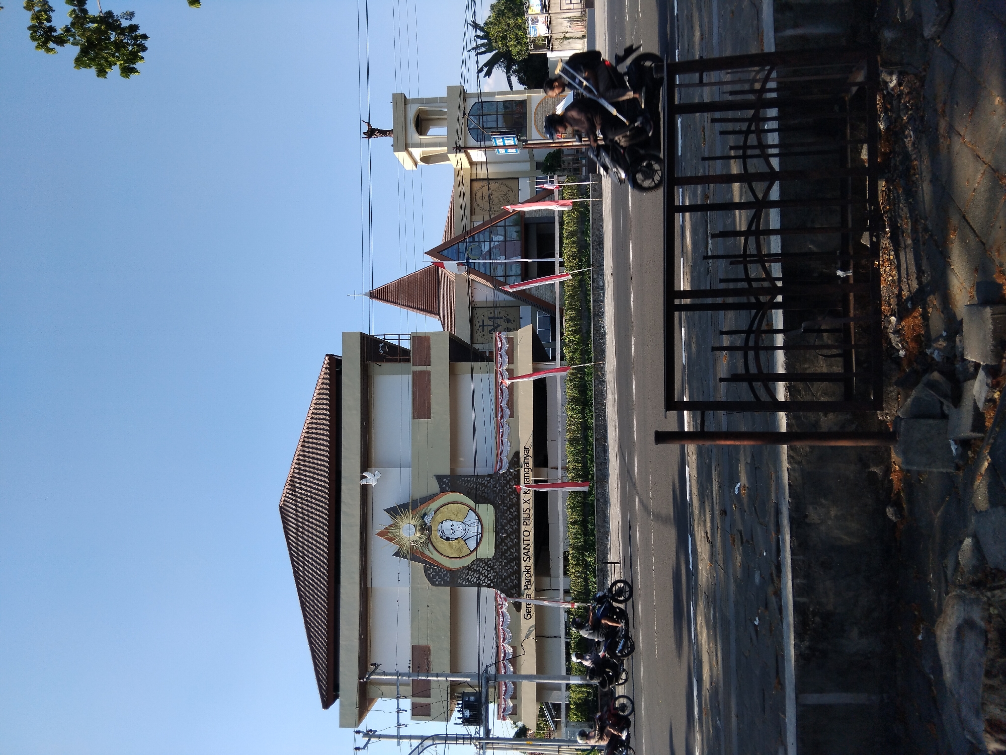 The image depicts the Paroki Santo Pius X church in Karanganyar, Indonesia, under a clear blue sky. The church features a distinctive modern architectural design with a prominent pitched roof and a central stained-glass facade. A statue of Jesus is visible atop a bell tower to the right of the main building. In front of the church, a landscaped area separates it from a road where several motorcycles are traveling. One motorcycle has two occupants, while others appear to be ridden by single individuals. The road surface is asphalt with white lane markings. A dark metal fence is visible in the foreground, bordering a concrete wall and debris-strewn ground. The overall scene captures a segment of urban life with religious architecture and daily transportation.