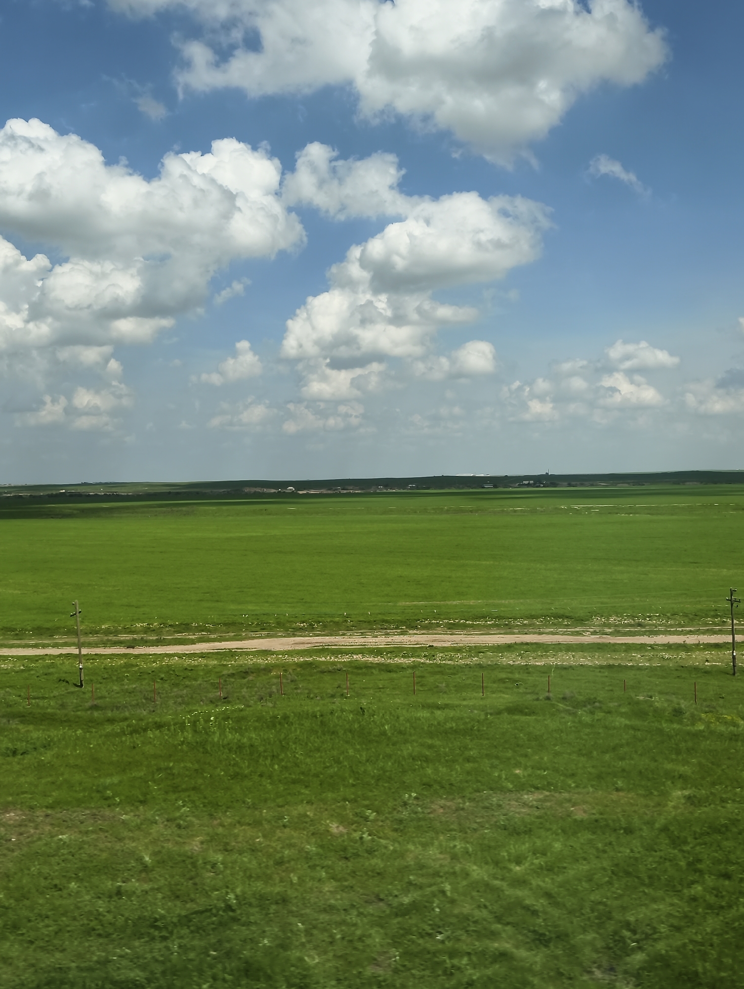 The image depicts a wide, green landscape under a bright sky with scattered white clouds. In the distance, a faint treeline is visible. A dirt road cuts horizontally across the midground of the scene. In the foreground, there is a grassy area with sparse vegetation and a fence of thin red posts. There are also telephone poles. The location is Unknown, Unknown.