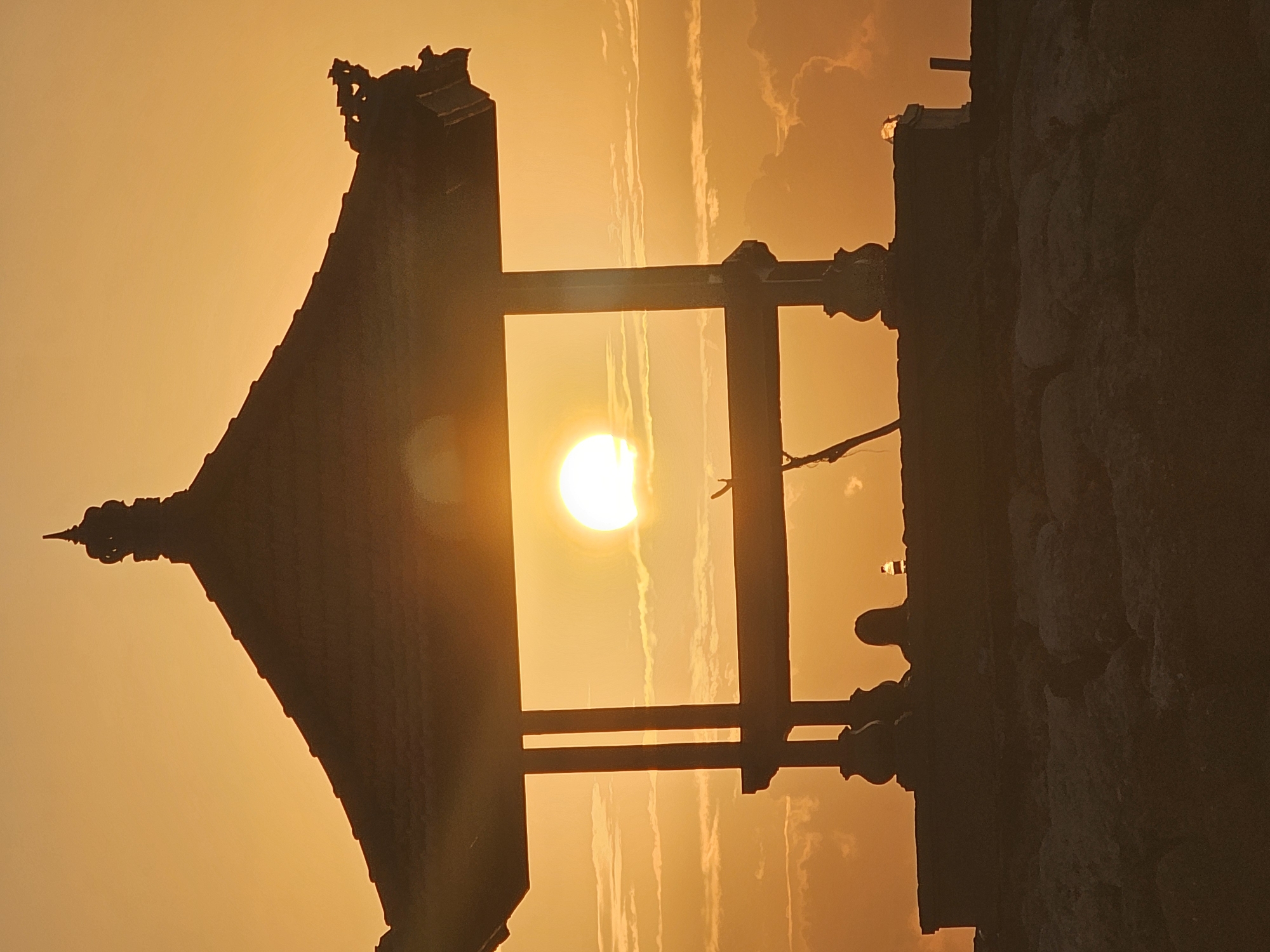 A silhouette of a traditional Balinese pavilion is positioned in the foreground, framing a brilliant sunset. The pavilion features intricate architectural details on its roof and pillars. Through the open structure, the sun is a bright, glowing orb against a sky filled with soft, wispy clouds, all bathed in a warm, golden-orange hue. Below the pavilion, the silhouette of a person can be seen, suggesting a solitary moment of contemplation or appreciation of the view. The base of the pavilion rests on a foundation of rough, dark stones. The overall scene evokes a sense of tranquility and natural beauty, characteristic of coastal sunsets in Bali, Indonesia. While the exact location is not specified, this scene is highly representative of the many beachside pavilions found in areas like Seminyak or Sanur in Denpasar.