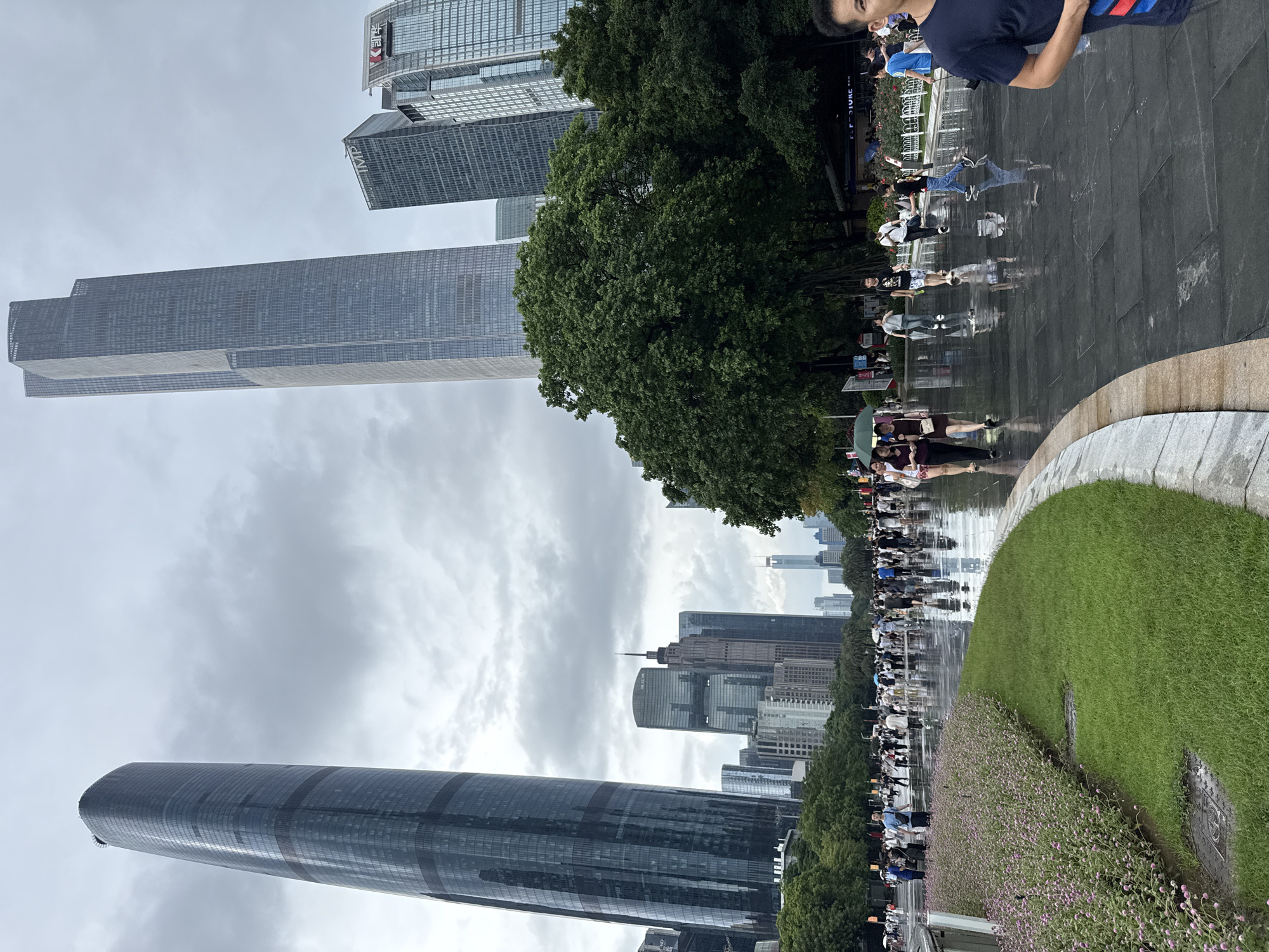 In Tianhe District, China, a wet, paved pedestrian walkway is filled with people walking under a cloudy sky. The ground is slick with rain, reflecting the surrounding modern skyscrapers. To the left, a tall, curved skyscraper with a dark, reflective facade dominates the skyline. In the middle ground, a dense cluster of green trees partially obscures a mix of taller and shorter buildings, including one with the "IMP" logo and another with Chinese characters. Further in the background, more skyscrapers rise against the overcast sky, some appearing thin and distant. A neatly manicured lawn bordered by a flower bed with small pink flowers curves along the left side of the walkway. People are dressed in casual attire, and some are holding umbrellas. The overall scene depicts a busy urban environment after a rainfall, with a blend of nature and modern architecture.