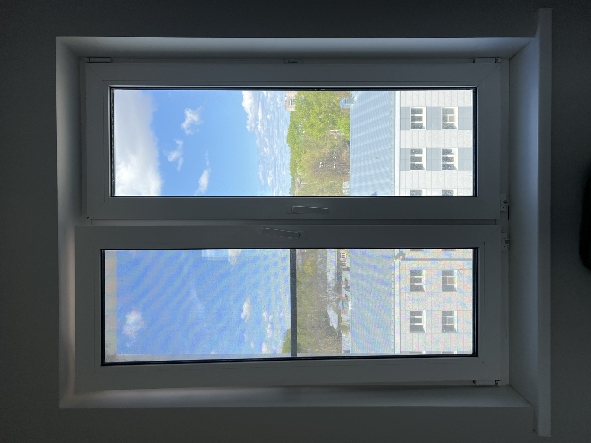 A low-angle, interior view of a closed, double-paned window in a likely residential setting. Outside the window, a view of a blue sky with scattered clouds can be seen. Also visible are trees with green foliage, indicating a springtime or summer setting, and a multi-story building with multiple windows. The building is light-colored. The image does not provide enough data to determine if this is Vologda, Russia. There are no visible people.