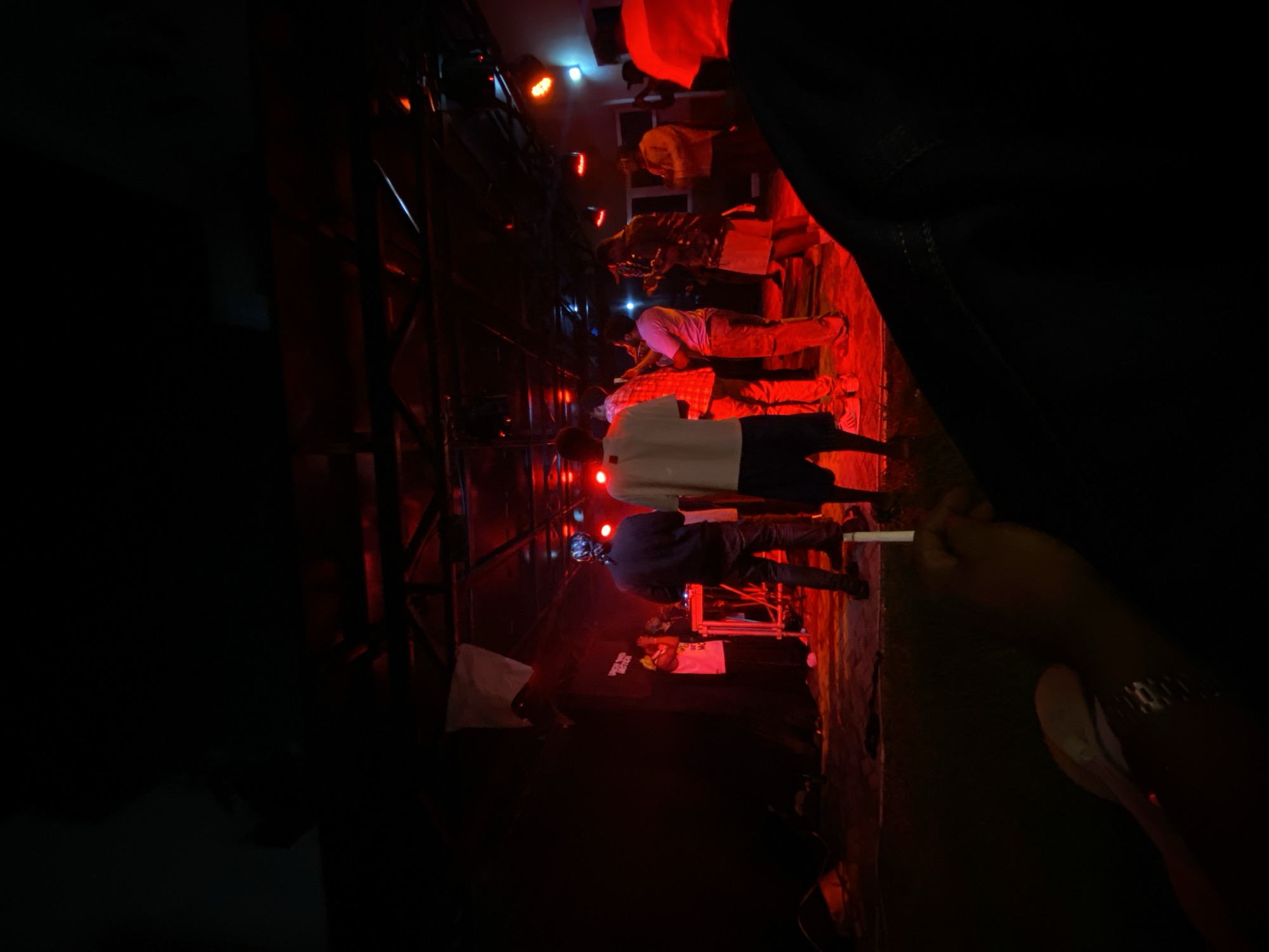 The image depicts a dimly lit indoor venue, likely a performance space, under red-toned lighting. A group of people are on a stage, some appearing to be performers, with musical equipment visible. The crowd is not clearly visible, but the perspective suggests a gathering of some sort. The presence of a cigarette in the foreground suggests a relaxed, possibly informal, social setting. Given the context of similar venues and performances, this scene is likely located in a vibrant entertainment district within Abuja, Nigeria.