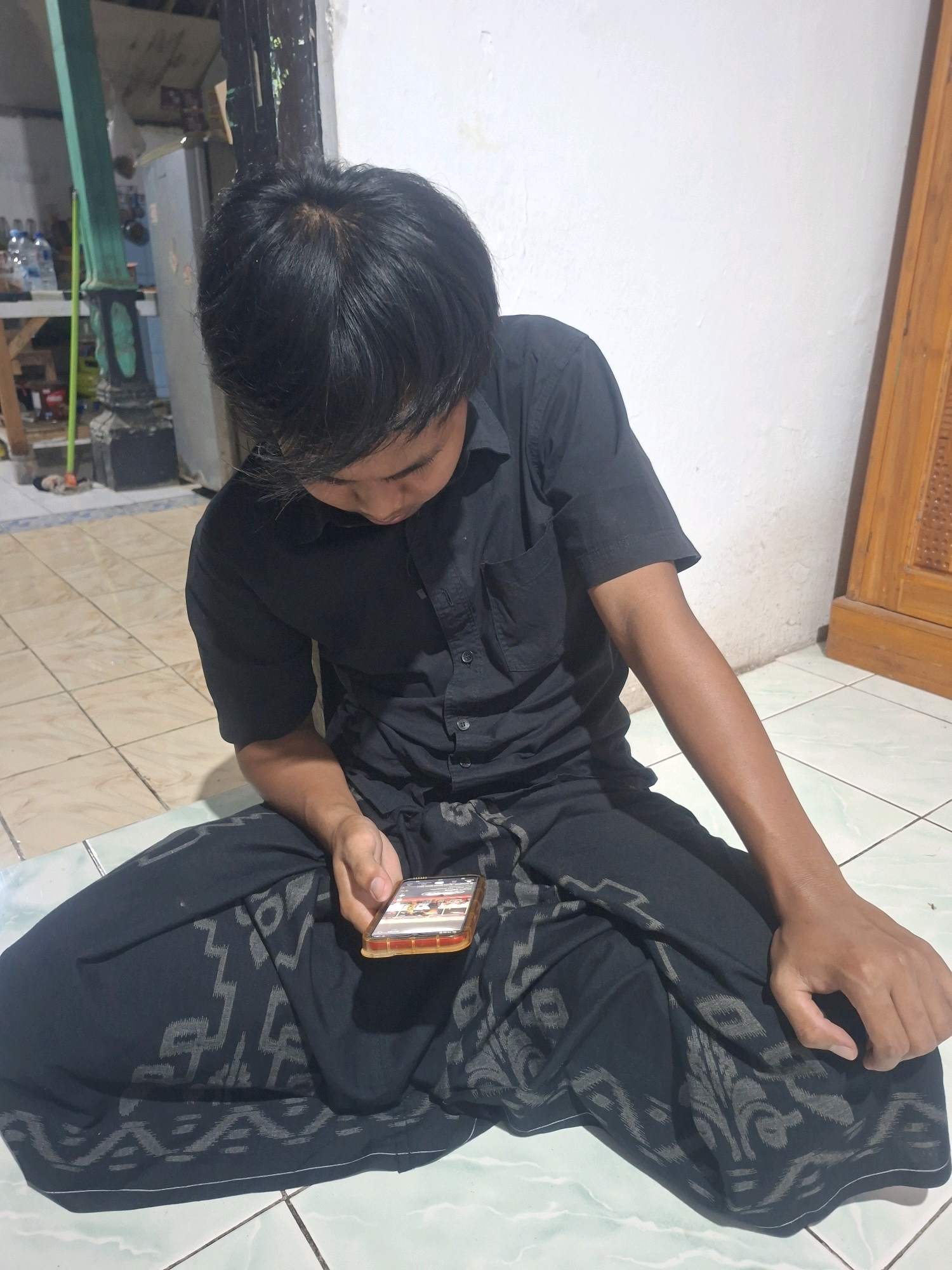 A young man wearing a black short-sleeved shirt and a dark patterned sarong sits on a tiled floor, looking at a smartphone in his hand. He is indoors, with a white wall behind him and a wooden door on the right. In the background, a refrigerator and shelves with items are visible. The floor is light-colored tile with a greenish-blue pattern. The image appears to be taken in a residential setting in Tongaswetan, Indonesia, possibly a home or community space.