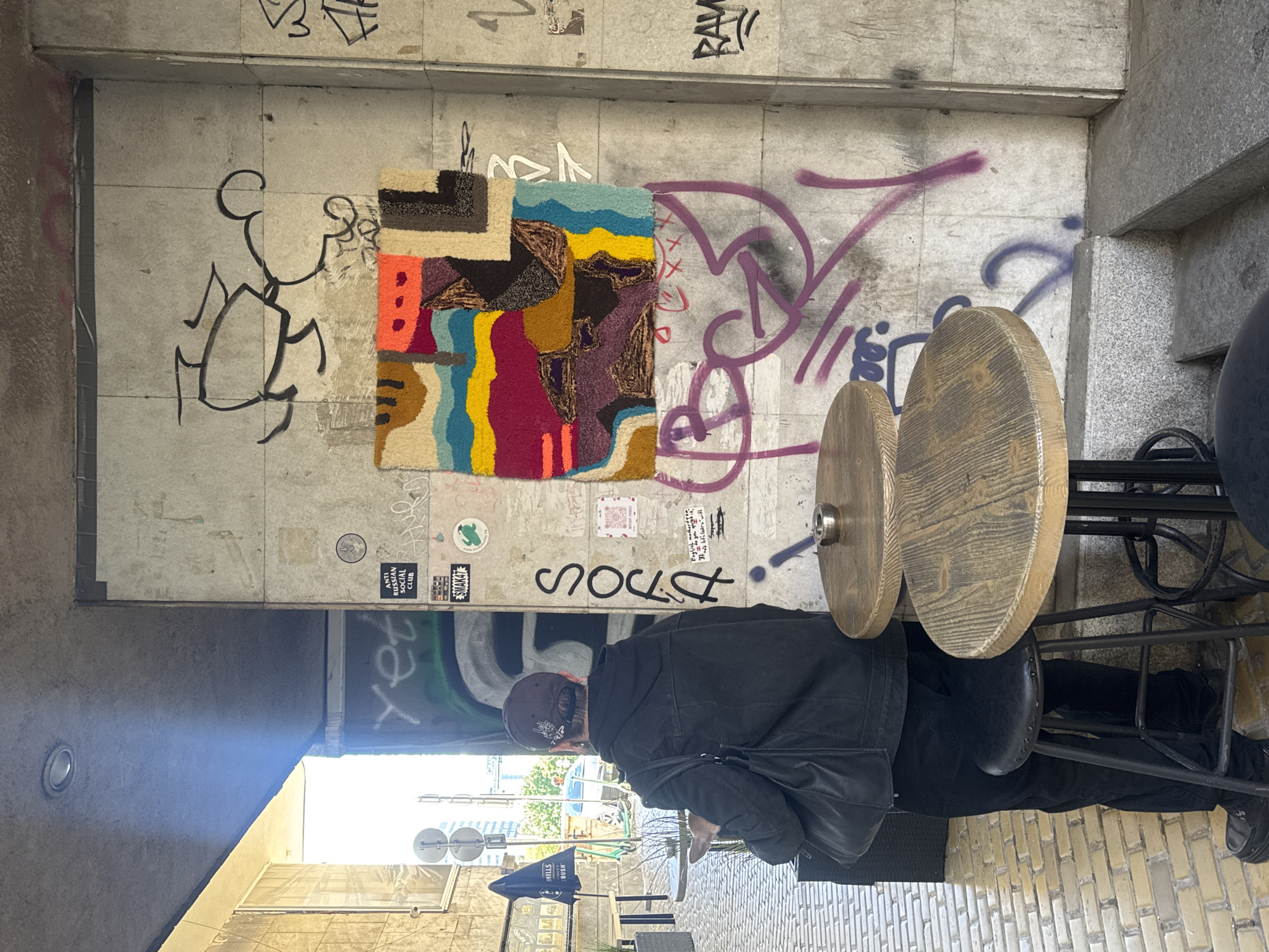 The image depicts an individual seated at a cafe table located against a graffiti-covered concrete wall in what appears to be an urban setting in Sofia, Bulgaria. The wall behind them is adorned with various tags, stickers, and an abstract, colorful rug. The rug consists of geometric shapes and contrasting colors like blue, yellow, red, brown, and black. The cafe setup includes two round wooden tables supported by a black metal frame. The person is wearing a dark jacket, a baseball cap, and black pants. They are turned away from the camera, and a black shoulder bag is visible. The background shows a glimpse of a street with buildings, trees, and street furniture under clear daylight.