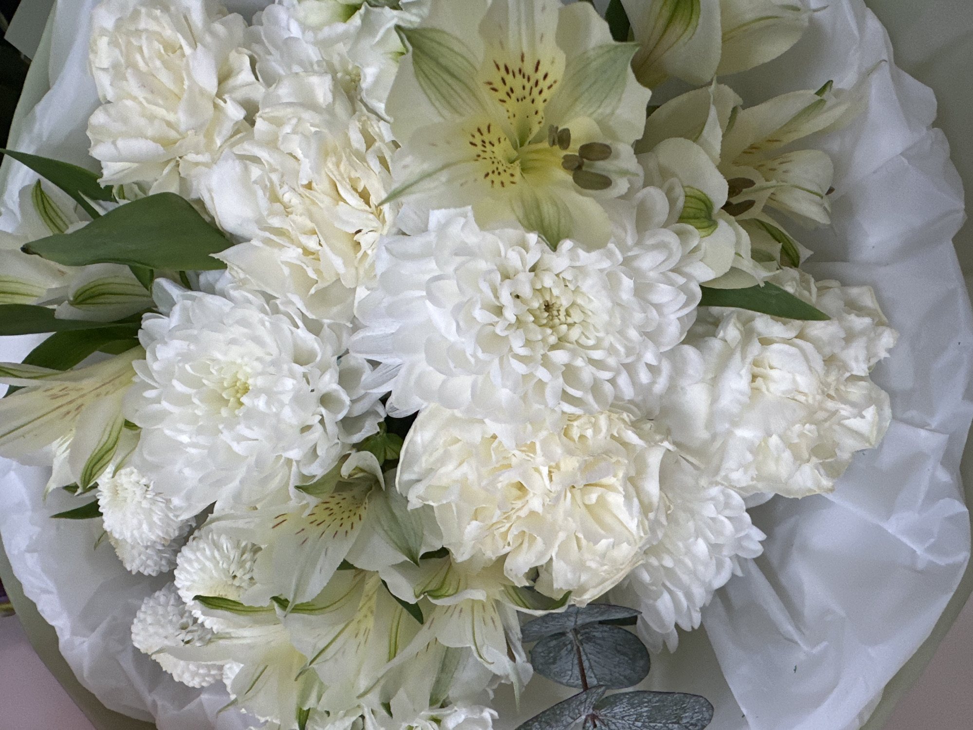 This image shows a bouquet of white flowers, including carnations, chrysanthemums, and alstroemeria. The flowers are arranged in a dense cluster and are wrapped in white paper. The color palette is dominated by white and light green. There is no evidence to suggest this image was taken in Yekaterinburg, Russia, or contains any specific information related to that location. There are no people in the image.