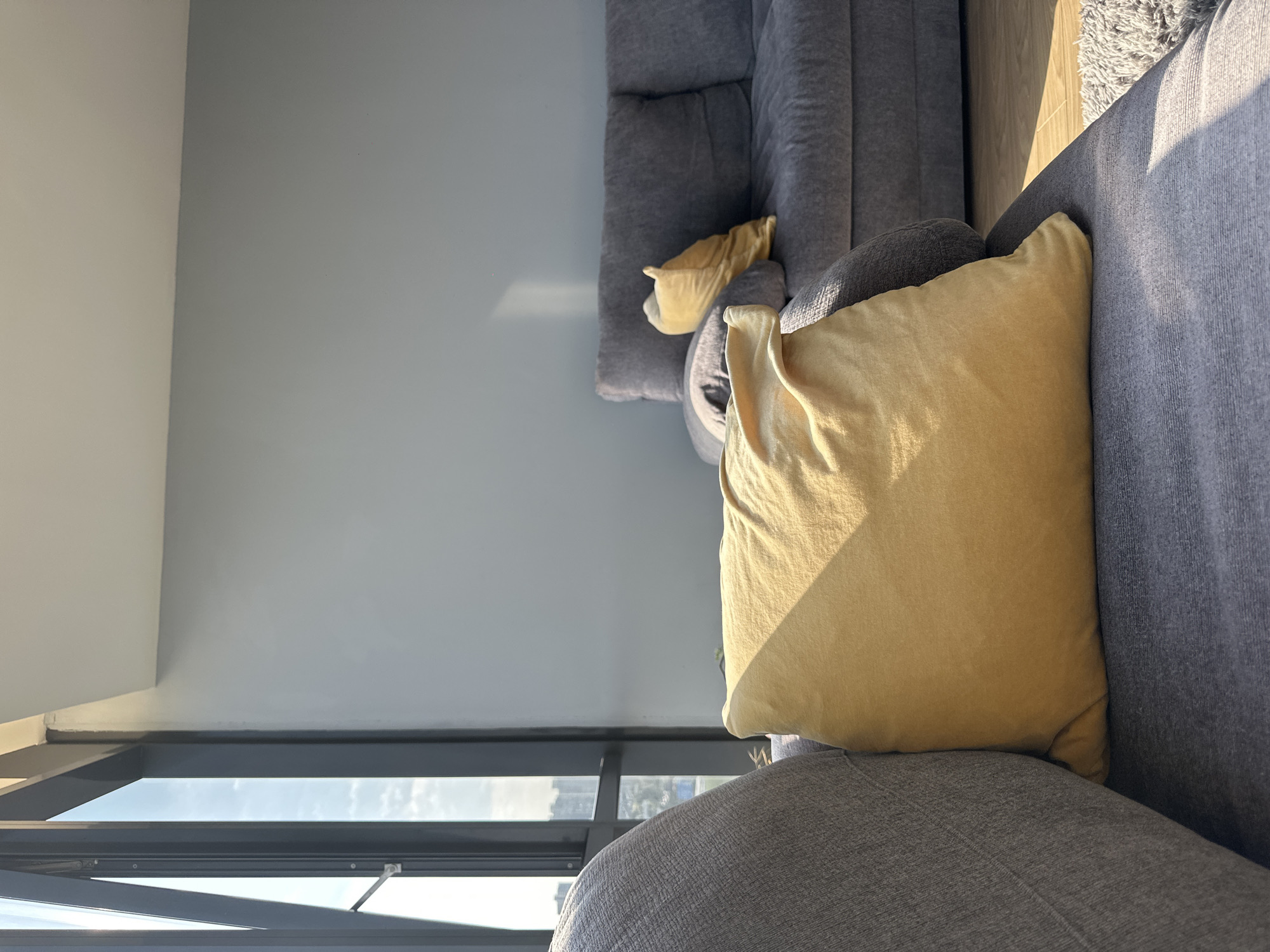 This image depicts a section of a living room interior. Two grey sofas are visible, one partially obscured and the other more prominently featured. The prominent sofa has a large, golden-colored pillow resting on it. There is also a smaller, similar-colored pillow on the partially obscured sofa. The scene is illuminated by natural light coming through a nearby window, which also offers a view of a cityscape. The location can be identified as Huixquilucan de Degollado, Mexico based on the buildings visible through the window. No people are present in the image.