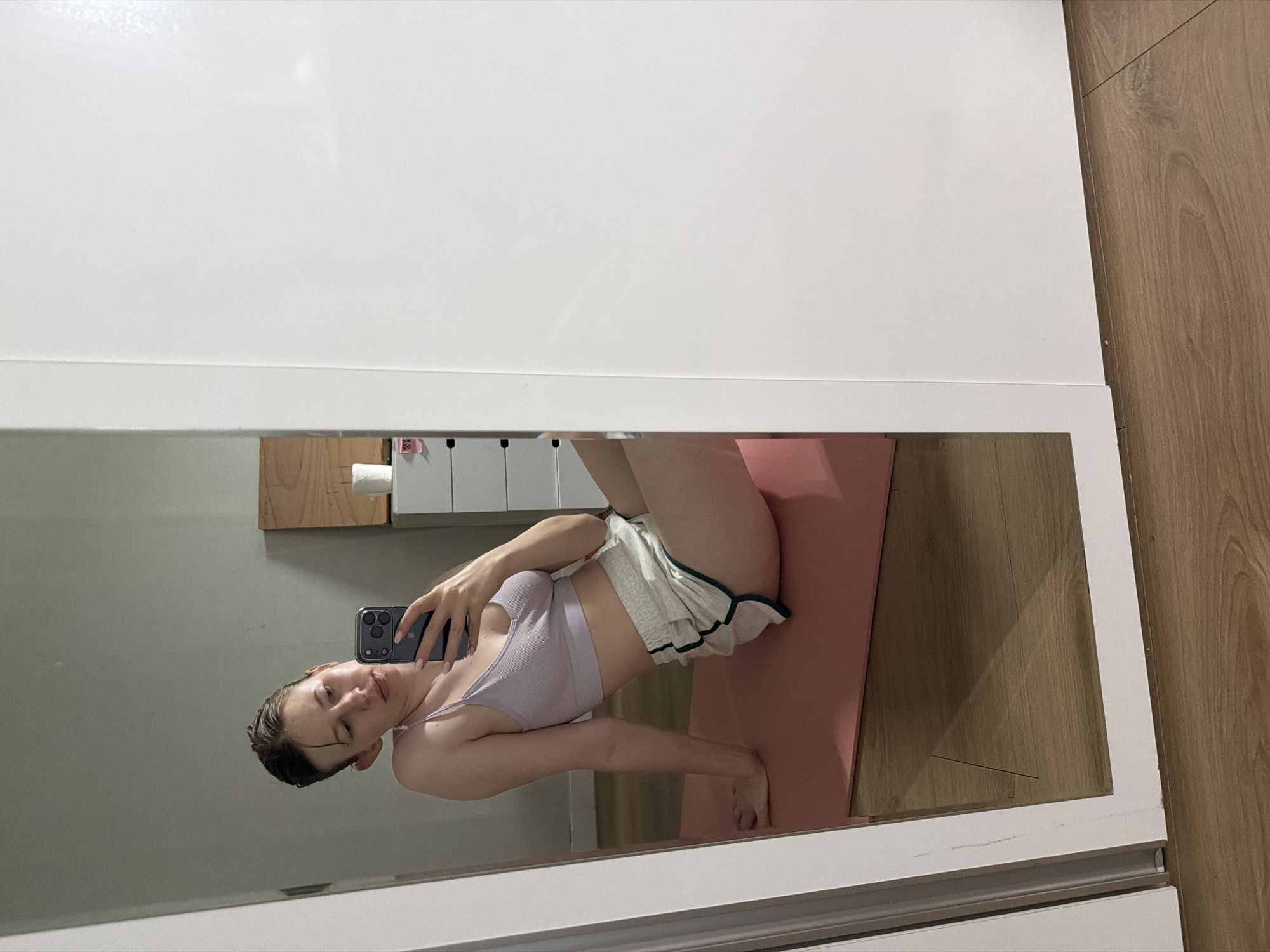A young woman with short, slicked-back hair is taking a mirror selfie. She is wearing a light purple ribbed sports bra and white shorts with green trim. She is sitting on a pink yoga mat on a wooden floor. The background shows a grey wall and a white closet. The image was taken in a room, likely a bedroom or workout space.