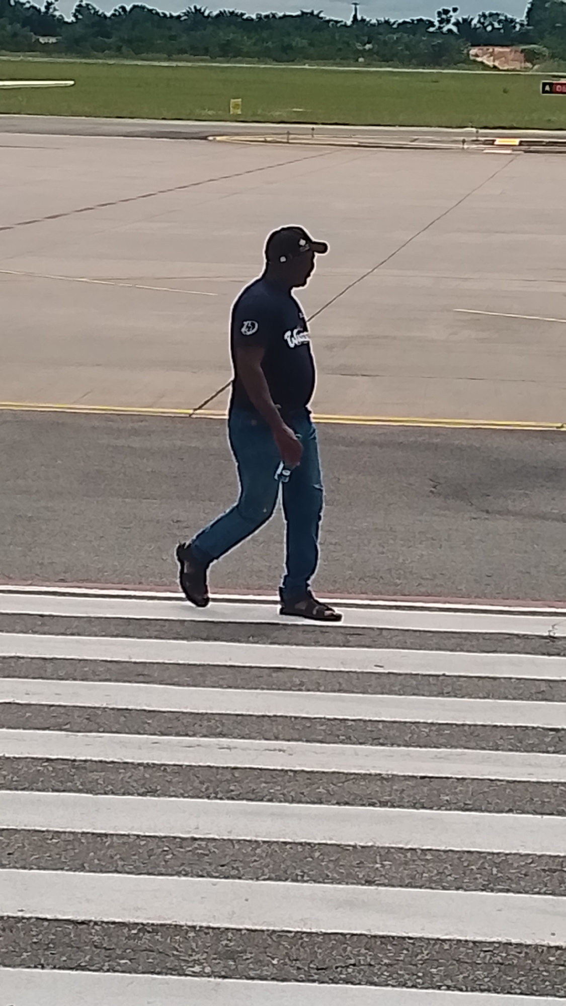 A man wearing a dark blue t-shirt, blue jeans, and sandals is walking across a pedestrian crossing on an asphalt surface. He is also wearing a dark baseball cap and appears to have earbuds in. He is holding a light-colored object in his right hand, possibly a phone or a bottle. The pedestrian crossing has wide white stripes. In the background, there is a green field with trees and vegetation visible, suggesting an outdoor location. The overall lighting indicates it is daytime, with a bright overcast sky. There is no clear indication of the weather, but it appears to be a neutral or slightly cloudy day. The scene is set at an airport or a similar large open area, given the asphalt and the expanse of the background. There is a yellow line on the asphalt to the left of the man. No visible text is discernible on the man's clothing or in the background. The man's posture suggests he is walking at a moderate pace.
