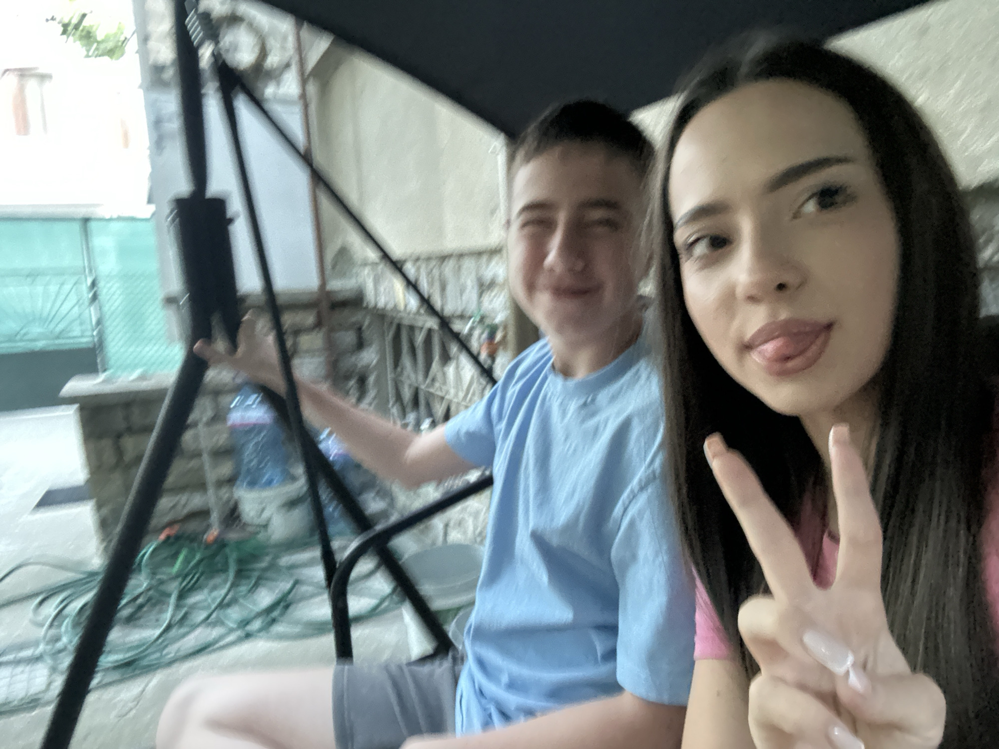 The image shows two individuals on a swing set, likely outdoors. A young man in a light blue t-shirt sits to the left, while a young woman with dark hair is to the right, making a peace sign and sticking out her tongue. The background reveals a building with a stone facade, a green hose, and various objects including a water bottle. The location appears to be a residential area, possibly in a backyard.