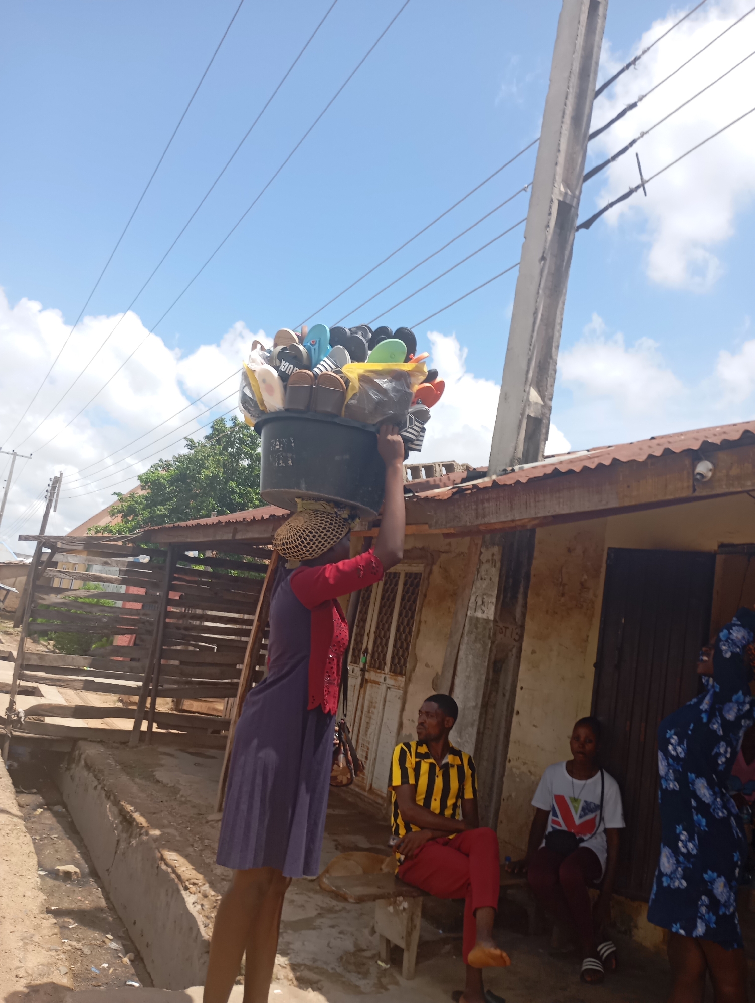 A woman balances a large black bucket filled with flip-flops on her head in a sunlit outdoor market scene in Shendam, Nigeria. She wears a purple dress and a straw hat. To her right, a person in a blue floral dress stands near a building with a corrugated metal roof. In front of the building, a man in a yellow and black striped shirt and red pants sits on a wooden bench, looking to his left. Beside him, a woman in a white shirt with a Union Jack design and dark pants also sits on a bench. The environment appears dusty with some debris on the ground and a concrete drainage ditch running alongside the path. Overhead, power lines crisscross a bright blue sky with scattered white clouds, and a utility pole stands to the right of the woman carrying the bucket.