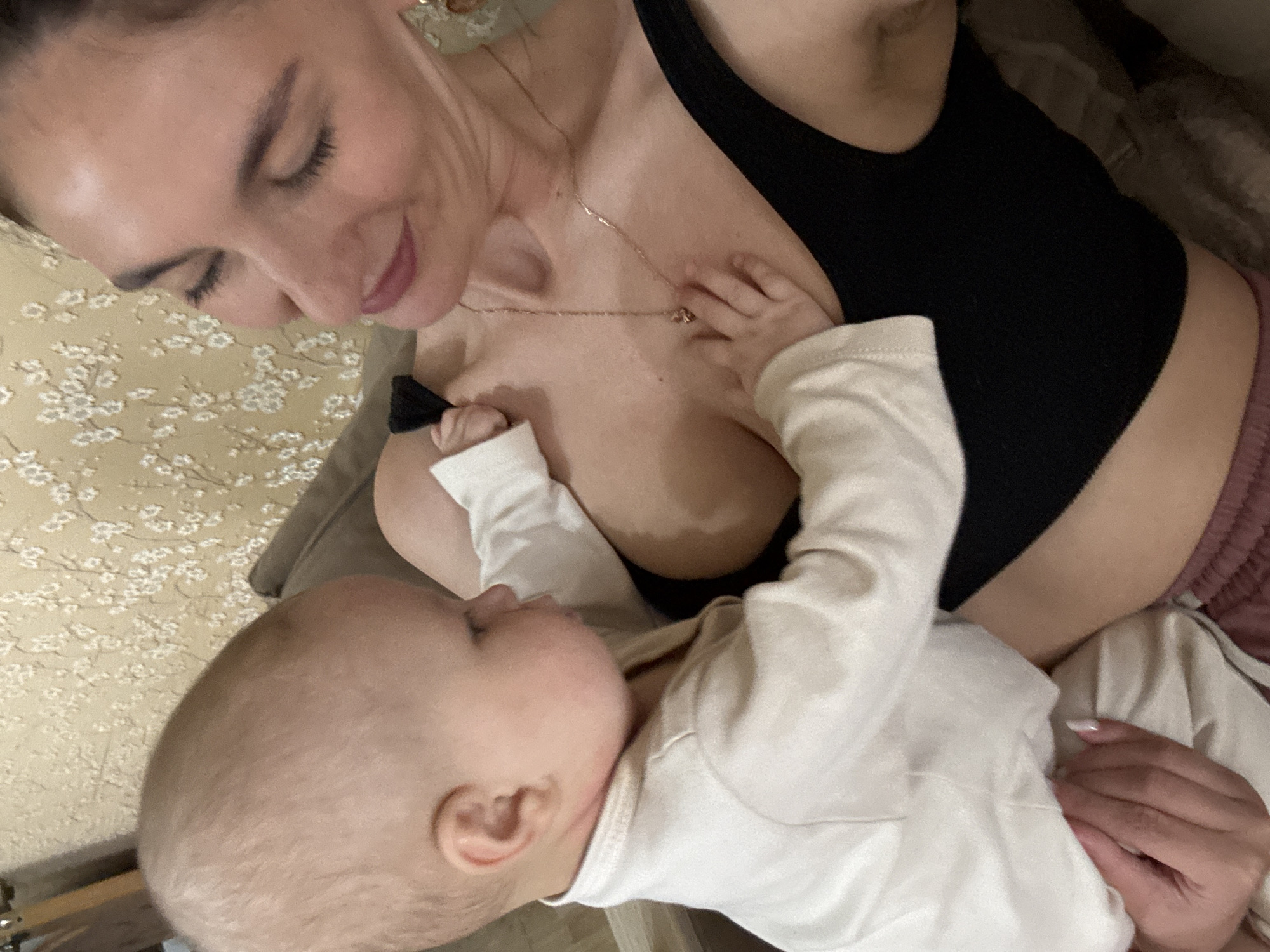 A woman with fair skin and dark hair is lying down, looking at a baby lying on her chest. The woman is wearing a black tank top and a gold necklace. The baby is wearing a white long-sleeved shirt. The background is a wall with a floral pattern and a light-colored sofa. There is no information to determine if the location is in the city of Sochi, Russia.
