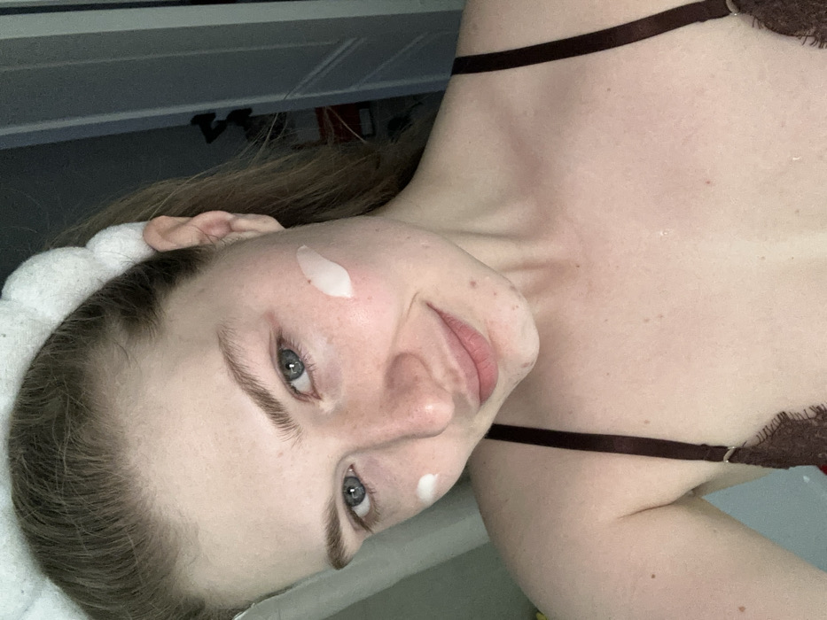 A fair-skinned person with brown hair and blue eyes poses for a close-up selfie, turned slightly to the side. A white headband holds back their hair. Dabs of white cream are visible on the right cheek and just below the chin. The person is wearing a brown bra with a thin strap. A white door or cabinet is visible in the blurred background.