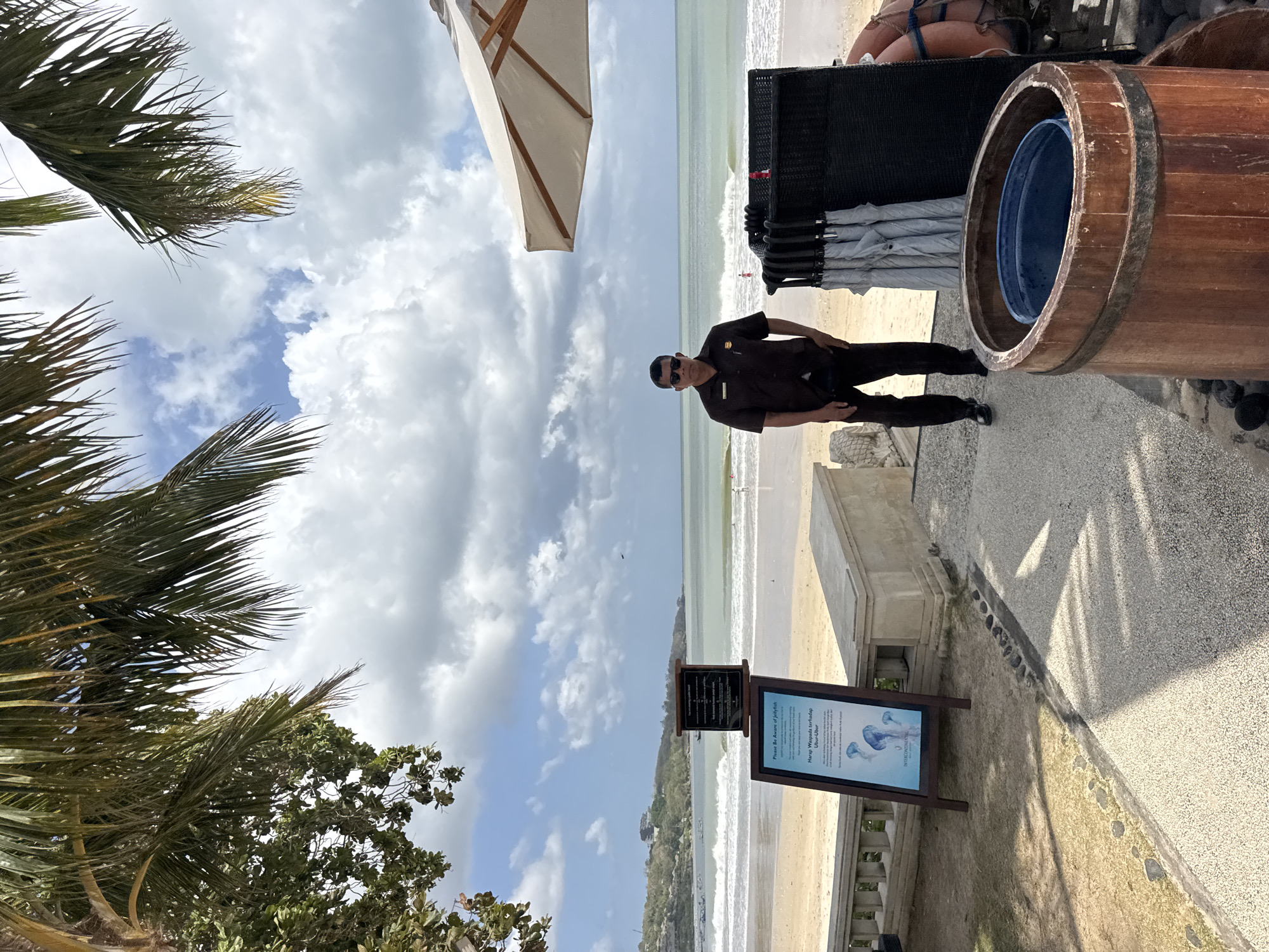 A person in a brown uniform stands on a pathway facing the ocean at a beach in Kuta Selatan, Indonesia. The person is wearing sunglasses and black pants. Behind the person are several closed umbrellas and a partial view of a beige umbrella. A wooden barrel with a blue bucket inside is in the foreground on the right. To the left of the person, a sign with images of jellyfish and text in English and Indonesian is displayed. The beach is visible in the background with waves breaking on the shore and a few people in the water. Palm trees with green fronds hang over the scene from the top left, and a clear sky with scattered clouds is visible.