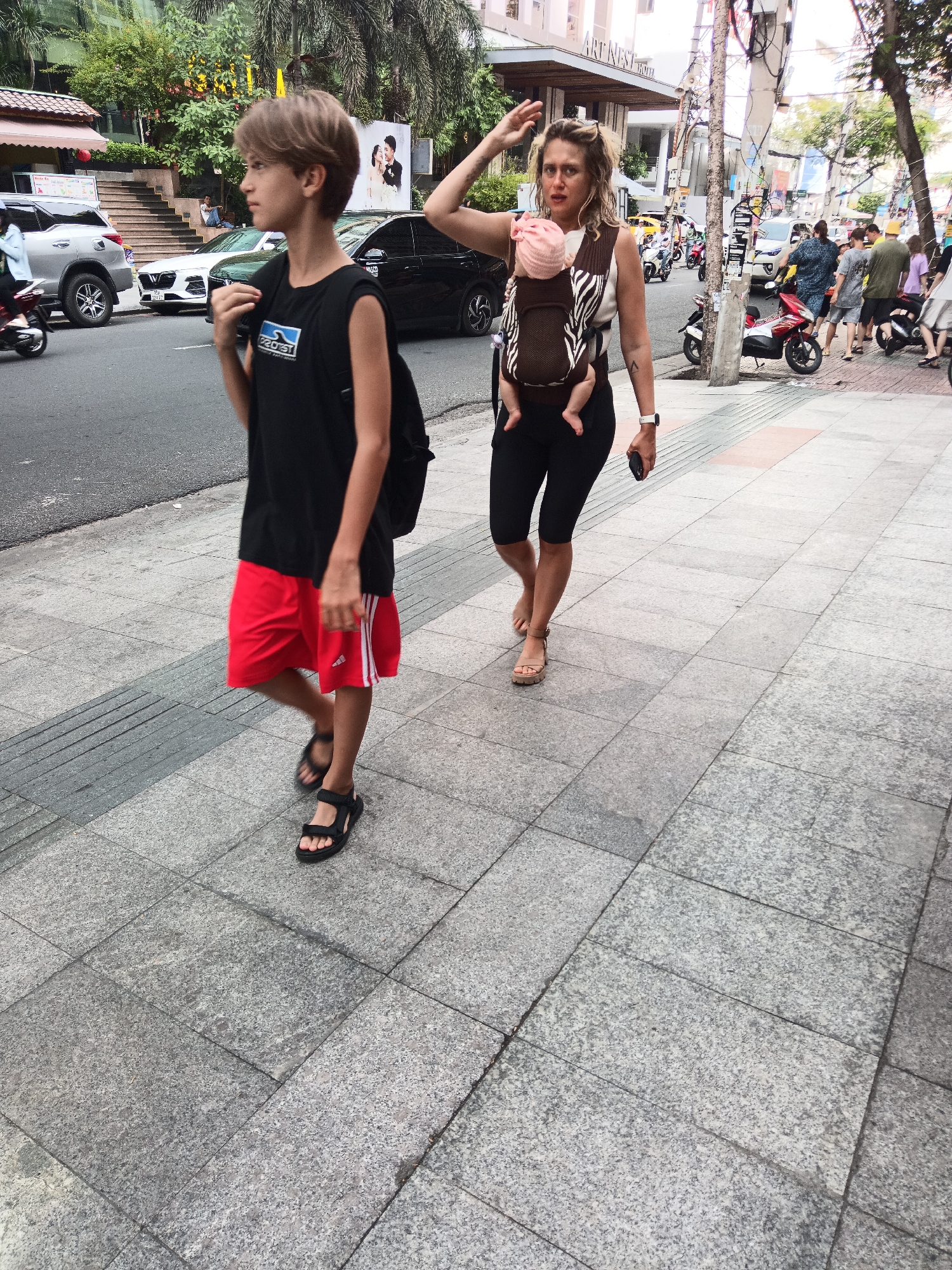 A young boy with light brown hair and a black t-shirt with a blue logo is walking on a sidewalk in Nha Trang, Vietnam. He is wearing red shorts and black sandals. Behind him, a woman is carrying a baby in a brown and white patterned baby carrier. She has blonde hair and is wearing black leggings and tan sandals. The background shows a street with cars, motorbikes, and buildings. The sidewalk is made of grey stone tiles.