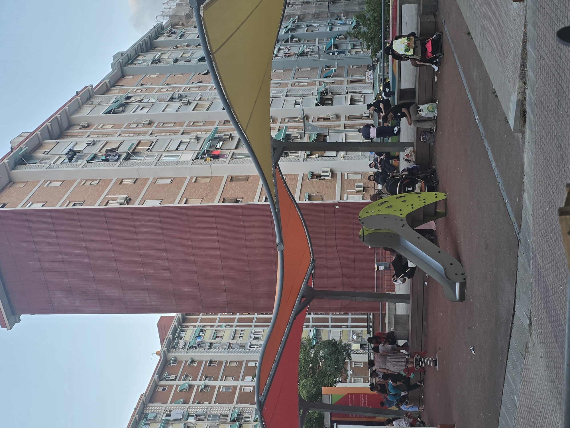 The image depicts a playground area in front of residential apartment buildings in Cornellà de Llobregat, Spain. The buildings are multi-story, with balconies and visible air conditioning units. In the foreground, a playground structure with a slide and a red spring rider is visible. Several people, including adults and children, are present in the playground area, some seated on benches, others standing or pushing strollers. Colorful fabric canopies extend over part of the playground, providing shade. The ground of the playground appears to be a dark, rubberized surface. Trash is scattered on the ground in the playground area.