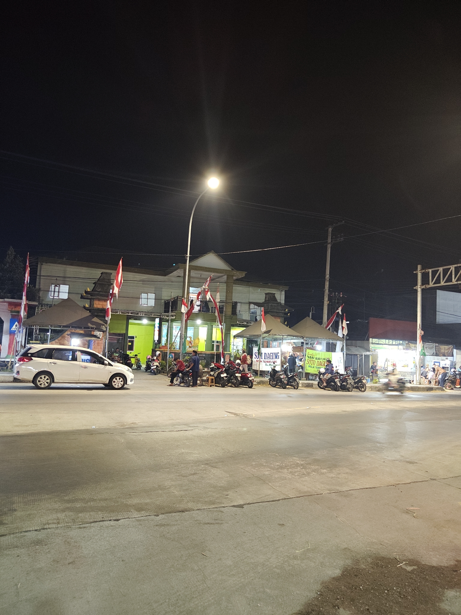 This is a nighttime photograph taken on a street in Bedah Lawak, Indonesia, showing a row of food stalls and a building. In the foreground, a white car is parked on the right side of the road. Several motorcycles are parked in front of the food stalls. The stalls are illuminated by lights, and banners display food items like "Soto Daging" and "Sate Ayam." People are seen standing and sitting at the stalls, engaged in eating and socializing. The background features a multi-story building with windows and flags. A street lamp illuminates the scene. The road surface is asphalt, and the overall atmosphere suggests a busy evening market or gathering.