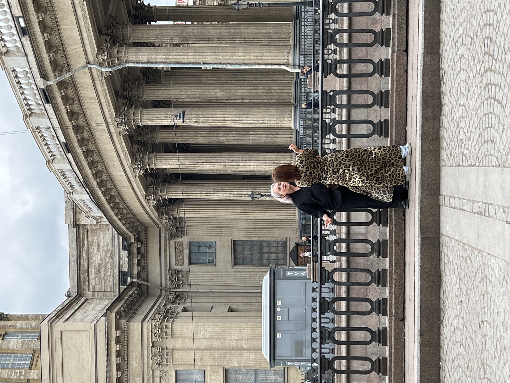 The image displays a street scene in Saint Petersburg, Russia, featuring the grand Kazan Cathedral with its distinctive curved colonnade. The monumental building, constructed from light-colored stone, dominates the left side of the frame, showcasing numerous tall, fluted columns with ornate Corinthian capitals and a richly decorated entablature. A metal drainpipe runs down one of the columns, and several windows are visible on the building's facade.
In the middle ground, a sturdy black metal railing with decorative oval-shaped links separates the building from a paved area in the foreground. Two women are prominently featured. The woman closer to the viewer, with light, swept-back hair, wears a black top and dark pants, leaning against the railing with a contemplative or serious expression. Behind her and slightly to her right, another woman wears a striking leopard-print coat. Her right arm is raised, possibly gesturing or holding a small object. In the background, behind the railing and among the columns, a few other people can be vaguely seen.
The foreground is paved with light-colored cobblestones or tiles arranged in a distinctive scallop or scale-like pattern. A grey portable toilet is situated behind the railing, positioned against the building's facade. The sky is overcast and grey, suggesting a cloudy day without direct sunlight.
Visible text on the portable toilet includes "TVALET" and "TOI" written vertically in white letters, and a horizontal "D" on its door. The overall atmosphere is quiet, possibly capturing a moment of observation or pause by the two main figures.
