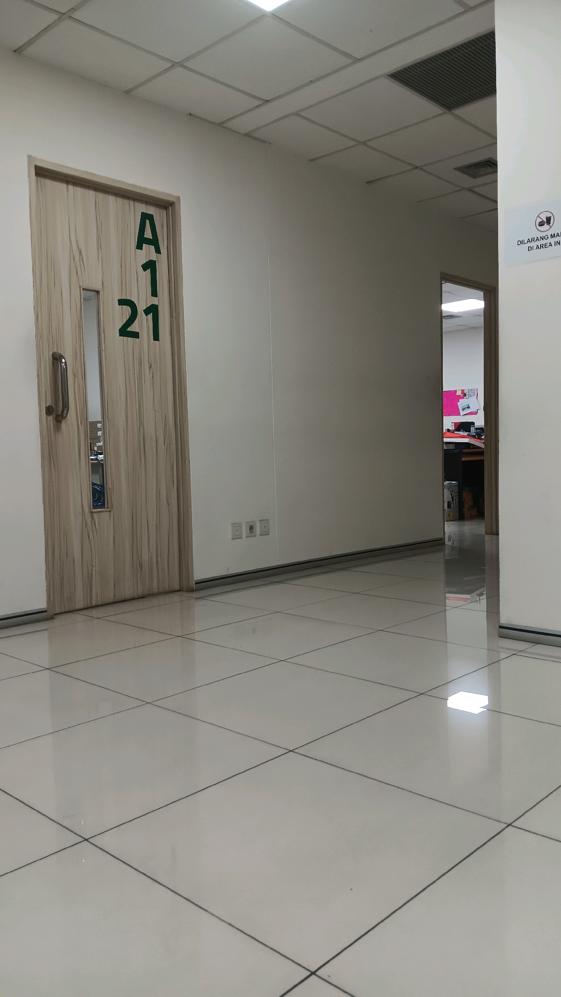 The image shows an interior hallway with a light-colored, tiled floor. On the left is a wooden door with a vertical window and a metal handle. The door is marked with the characters "A121" in green. To the right of the door, on the wall, are three electrical outlets. Further down the hallway, on the right wall, there is a sign with an image and text in Indonesian that reads "DILARANG MEROKOK DI AREA INI" (No smoking in this area). Beyond this, an open doorway reveals an office space with desks and cubicles. The ceiling is a grid of acoustic tiles with fluorescent lighting. The scene is a standard office or institutional building interior, likely within the city of Tangerang, Indonesia, given the Indonesian text on the sign. No people are visible in the image.