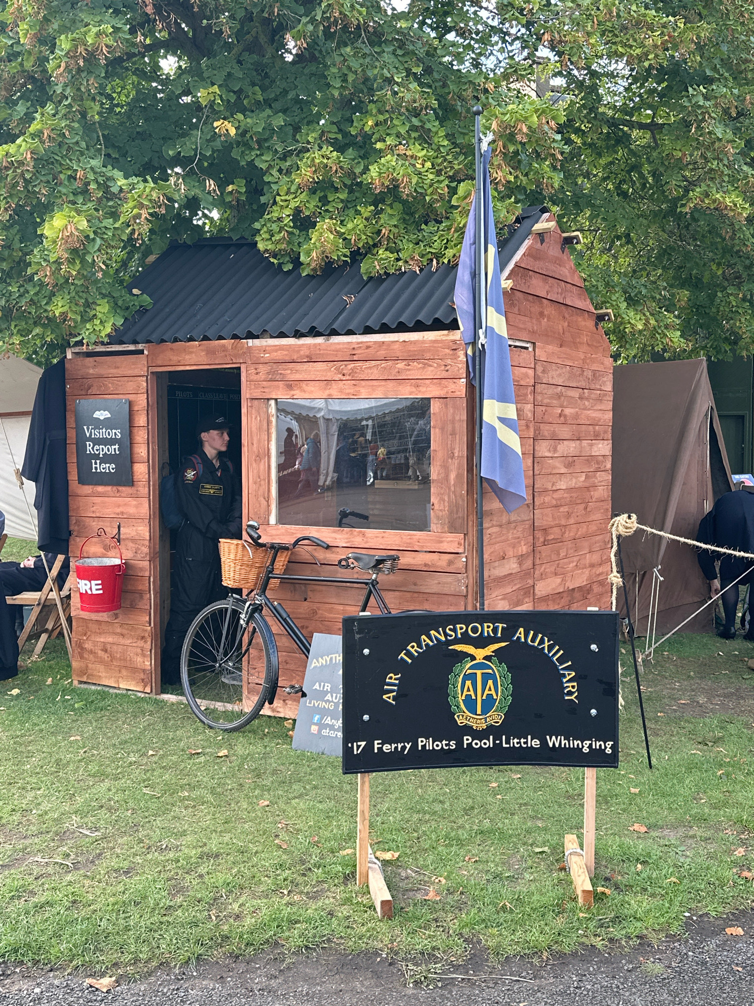 A wooden shed, likely a temporary structure, stands on a grassy area. A sign on the shed reads "Visitors Report Here." Inside the shed, a person in a dark uniform with a cap is standing, appearing to be on duty. A bicycle with a wicker basket is parked in front of the shed. A red bucket with "FIRE" written on it is hanging on the side of the shed. A flag, blue with yellow markings, flies from a pole attached to the shed. In the foreground, a black sign with text and a logo stands on wooden posts. The text on the sign reads "AIR TRANSPORT AUXILIARY" and below it, "Ferry Pilots Pool-Little Whinging." The surrounding area includes trees and other tents, suggesting an outdoor event or gathering. The scene is set in the city of South Cambridgeshire, United Kingdom.