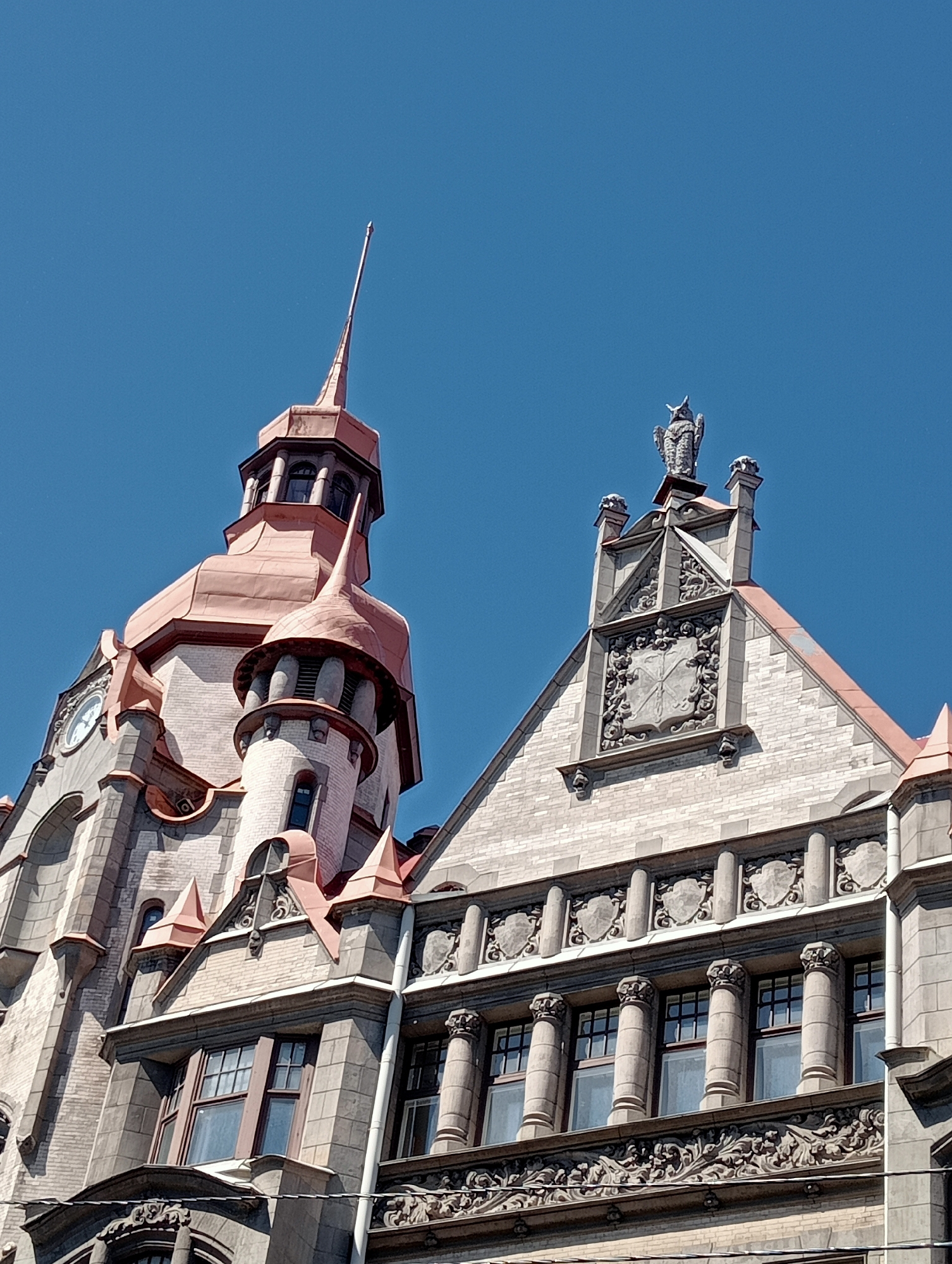 The image shows a close-up, low-angle view of the facade of a historic building in Saint Petersburg, Russia, under a clear blue sky. The architecture is elaborate and features a prominent tower with a copper-colored conical roof and a tall, slender spire. To the right of the tower is a gabled section of the building, also topped with a copper roof, adorned with a decorative crest featuring an owl sculpture. The facade itself is constructed with light-colored brick and stone, featuring intricate carvings, columns, and multiple windows. The left side of the building shows a circular clock face on the tower. The overall impression is one of grand, detailed historical architecture.